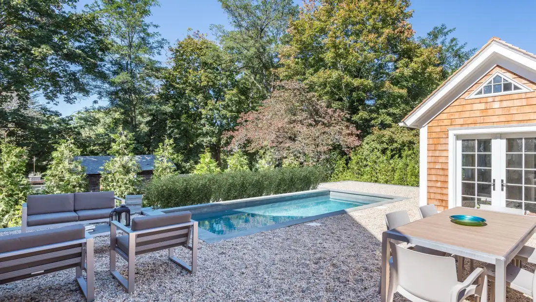 An outdoor pool beside a small building, with patio seating and leafy trees.