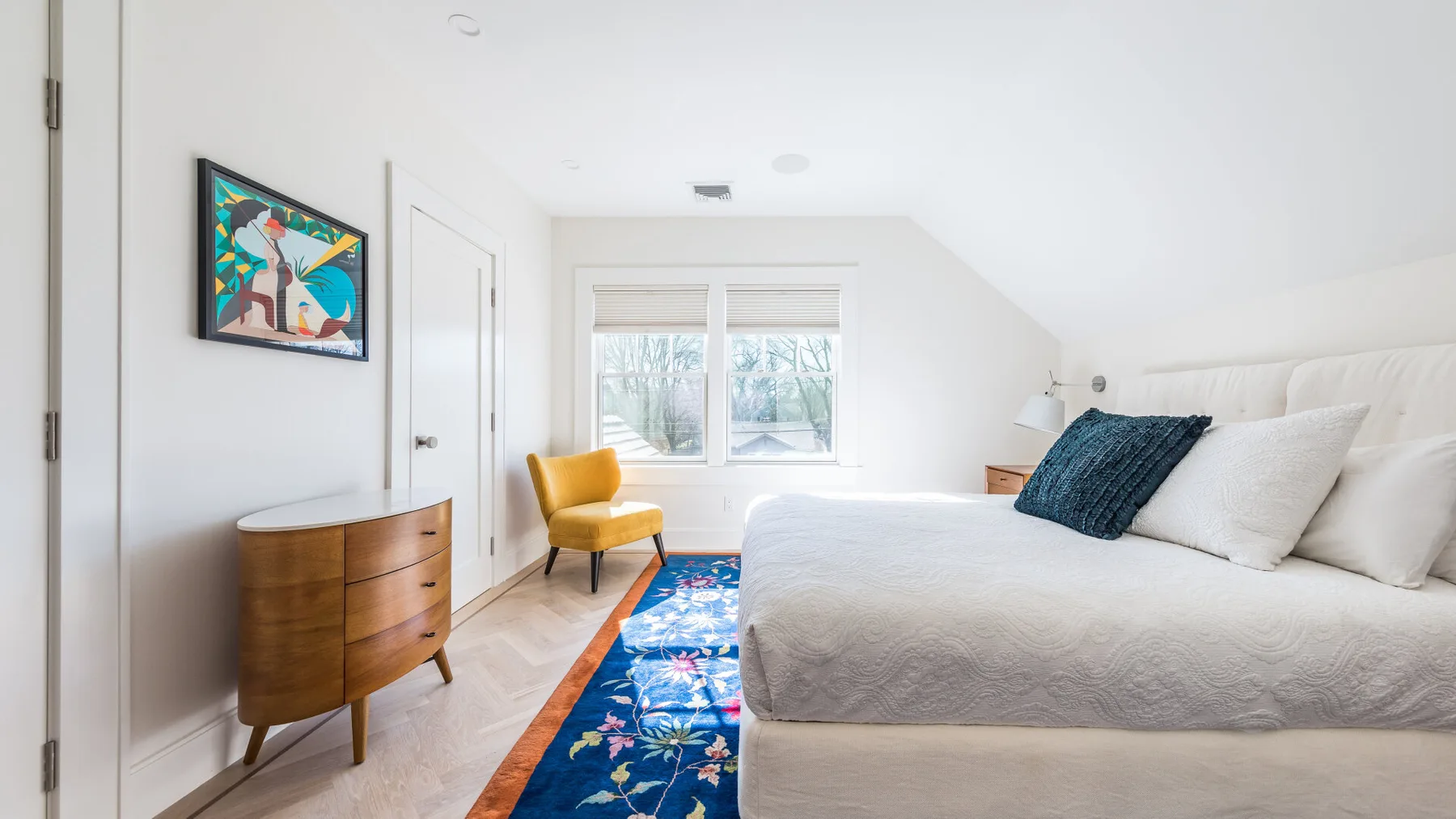 A bright bedroom with a neatly made bed, colorful rug, and wooden dresser.