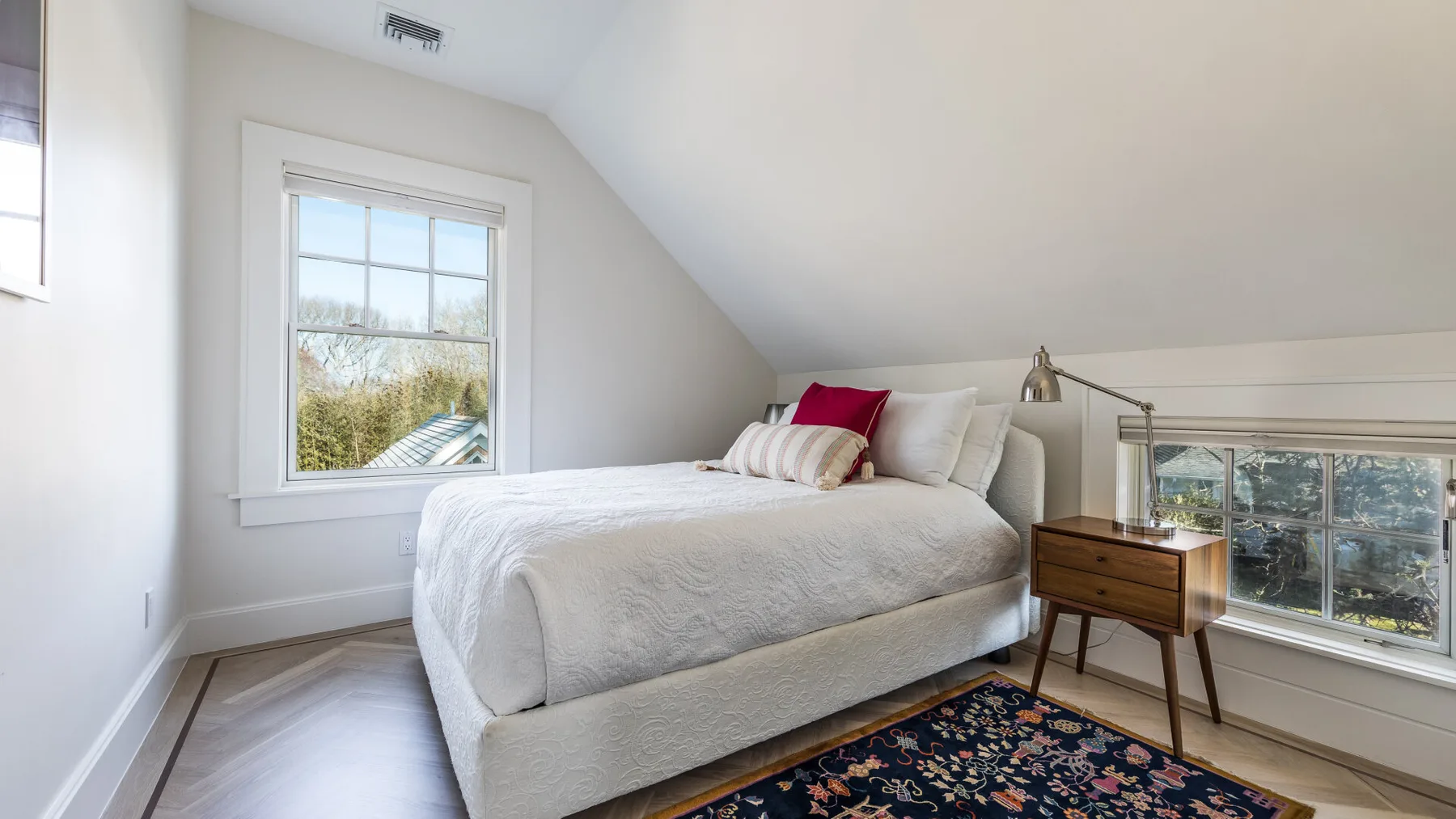 A bedroom with a bed, a patterned rug, and windows overlooking trees.