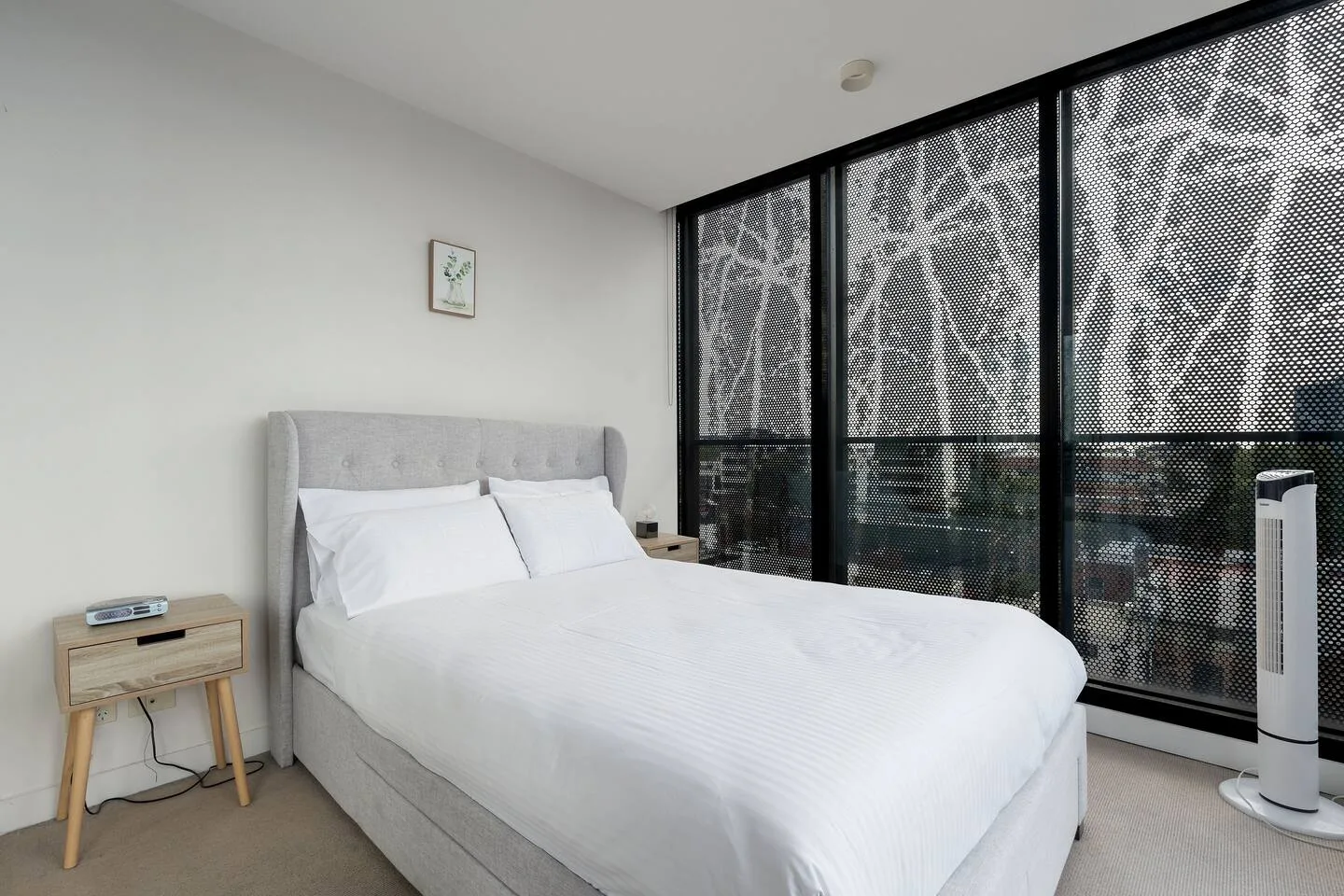 Un dormitorio con una cama doble junto a grandes ventanas y un ventilador de pie.