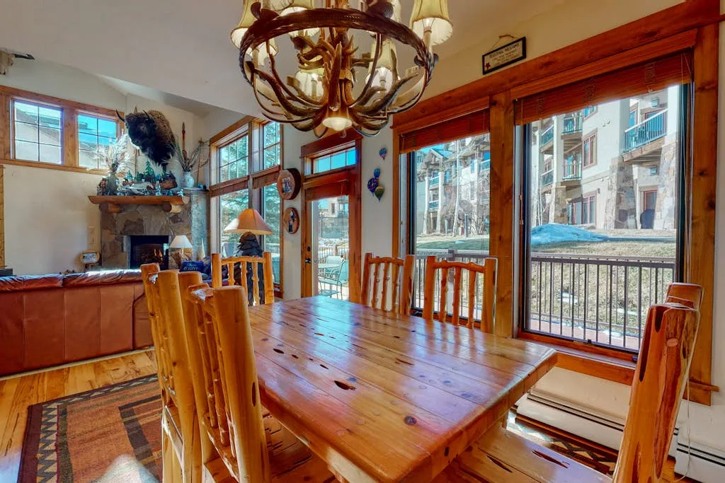 A wooden dining table and chairs sit beside large windows in a bright room.