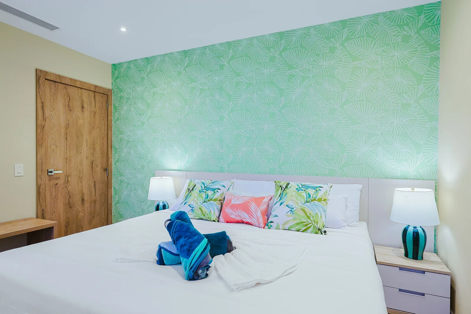 Rest easy on a plush bed with tropical-inspired linens and lively accent pillows against a refreshing floral wall.