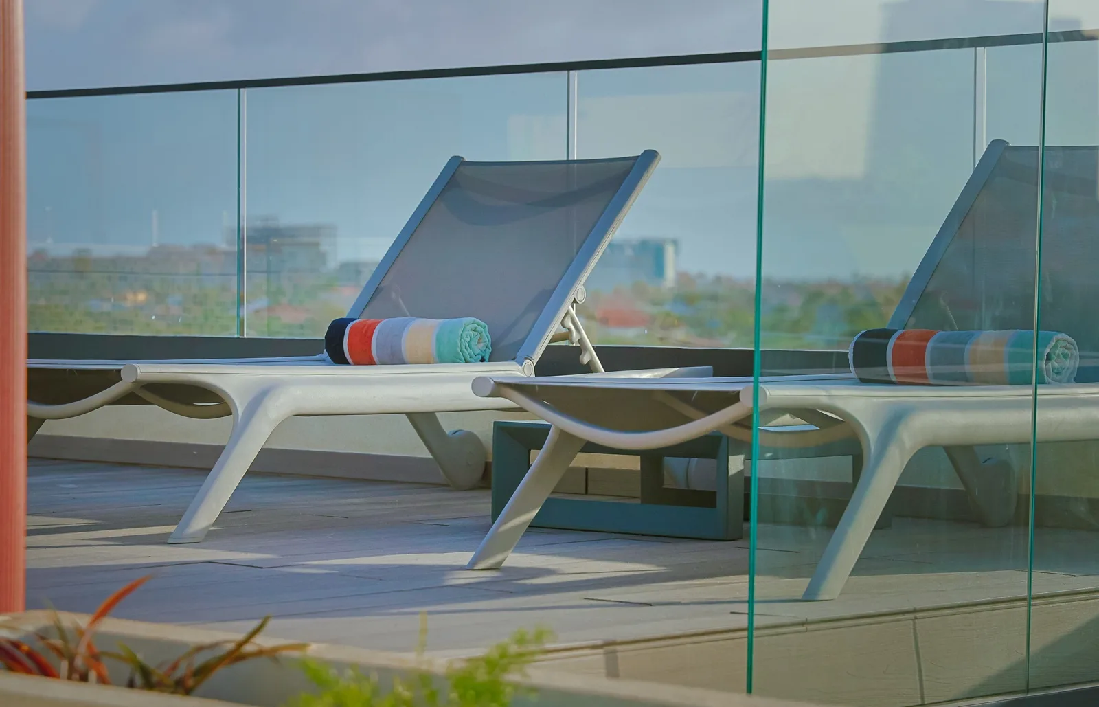 The rooftop sun loungers overlook the surrounding cityscape, offering a peaceful spot to unwind. Striped towels are ready for your comfort, and the glass railing frames the urban skyline beyond.