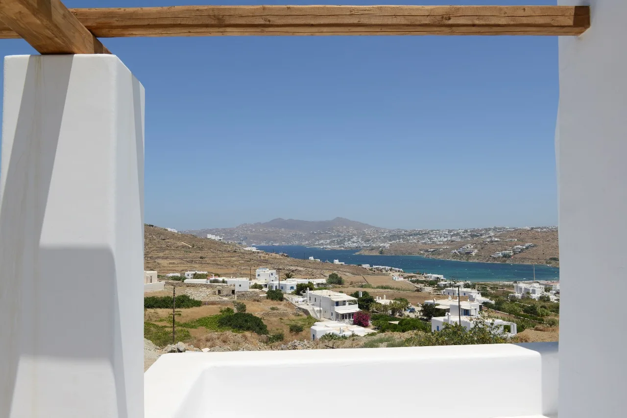 Una terraza blanca enmarca un paisaje costero con agua azul y colinas lejanas.