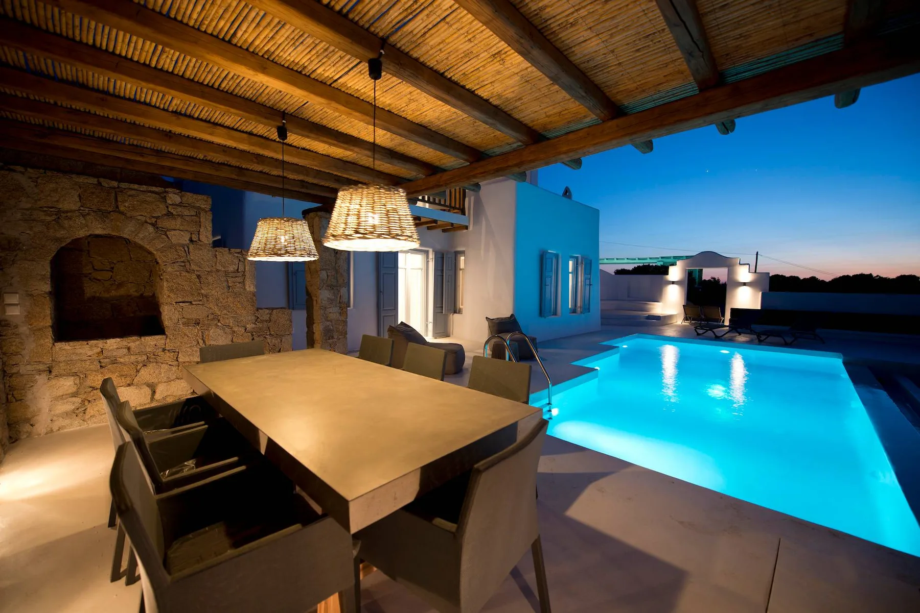 Comedor exterior junto a una piscina iluminada al anochecer.