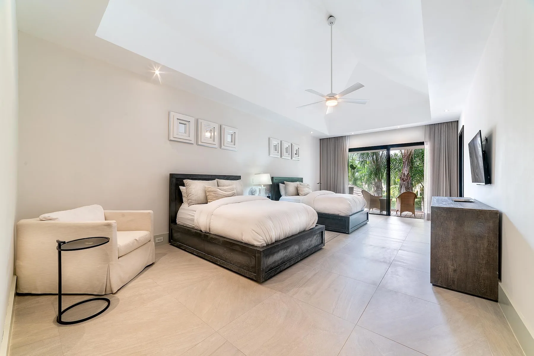 A bright bedroom with two beds, an armchair, and sliding glass doors.