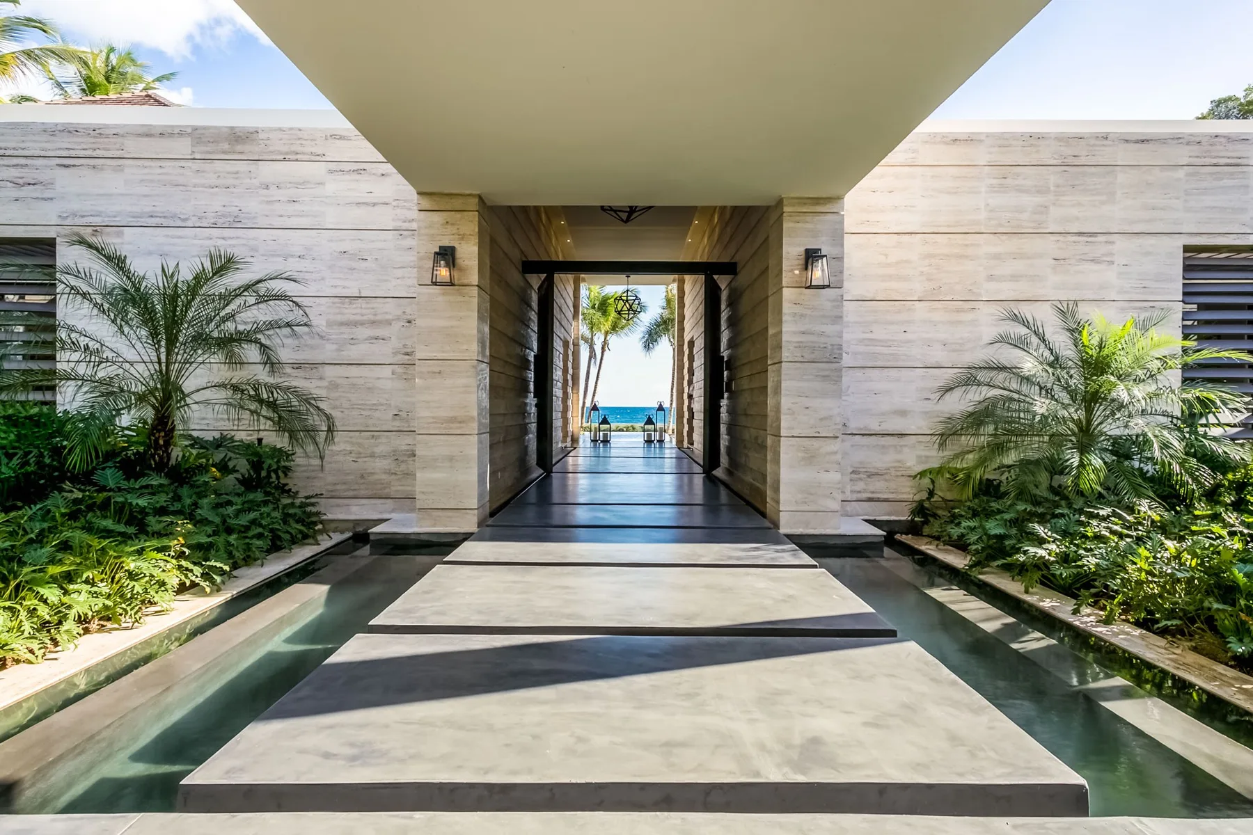 A covered entry walkway leads toward a bright outdoor view, with tropical plants along the sides.