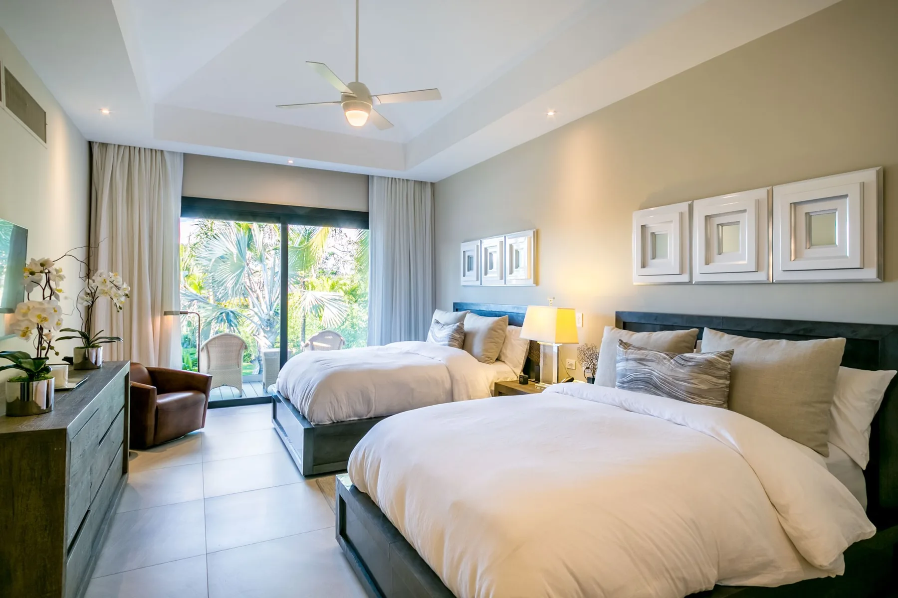 A bright bedroom with two beds, bedside lamps, and large windows overlooking greenery.
