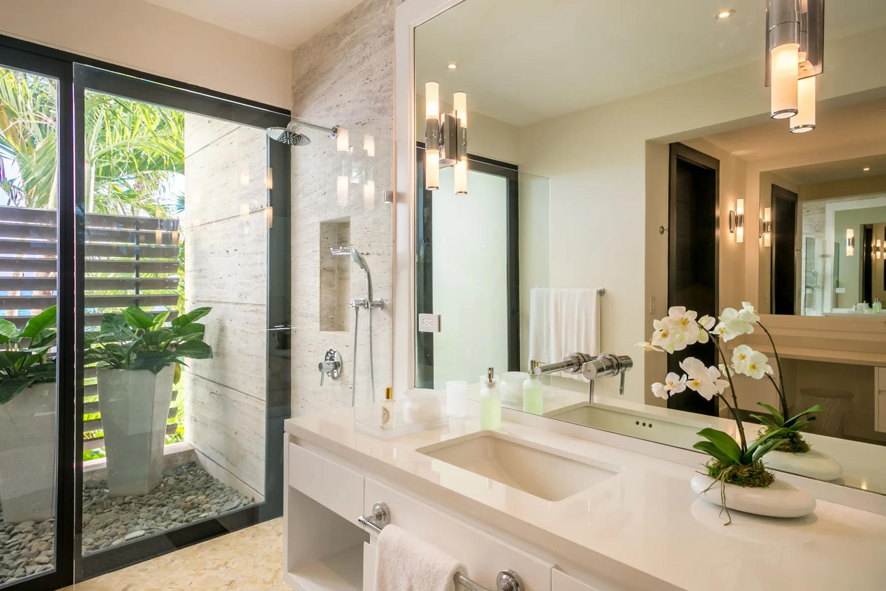 A bright bathroom has a broad vanity, mirror, and glass shower beside a garden view.