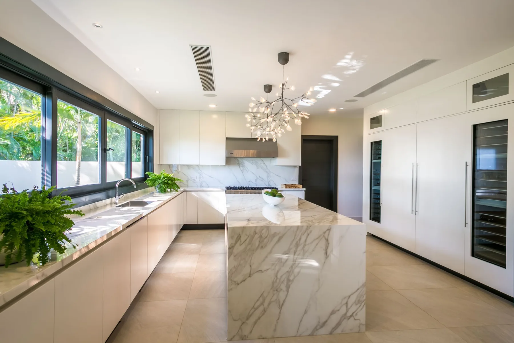 A bright modern kitchen has pale cabinetry, large windows, and a marble island.