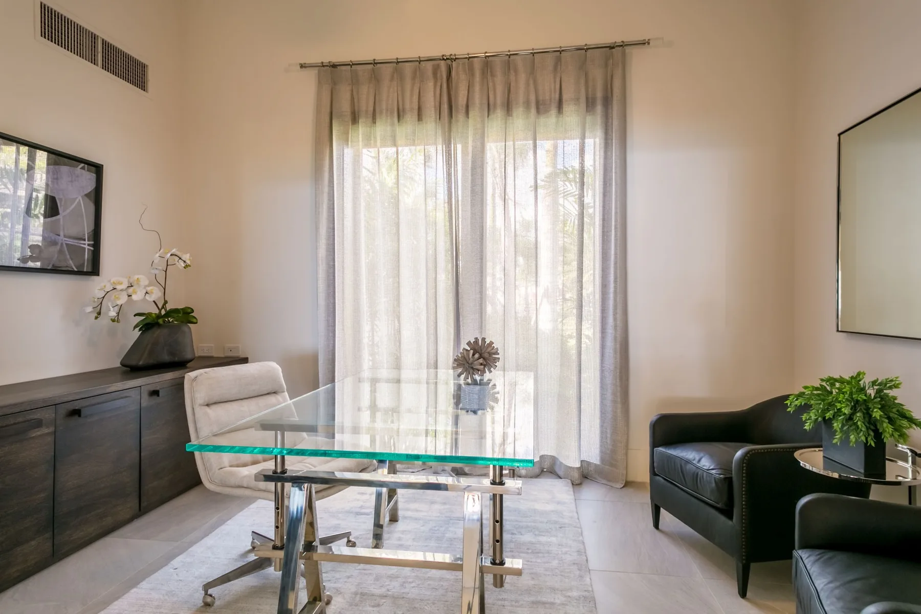 A dining area with a glass table and seating beside sheer curtains.