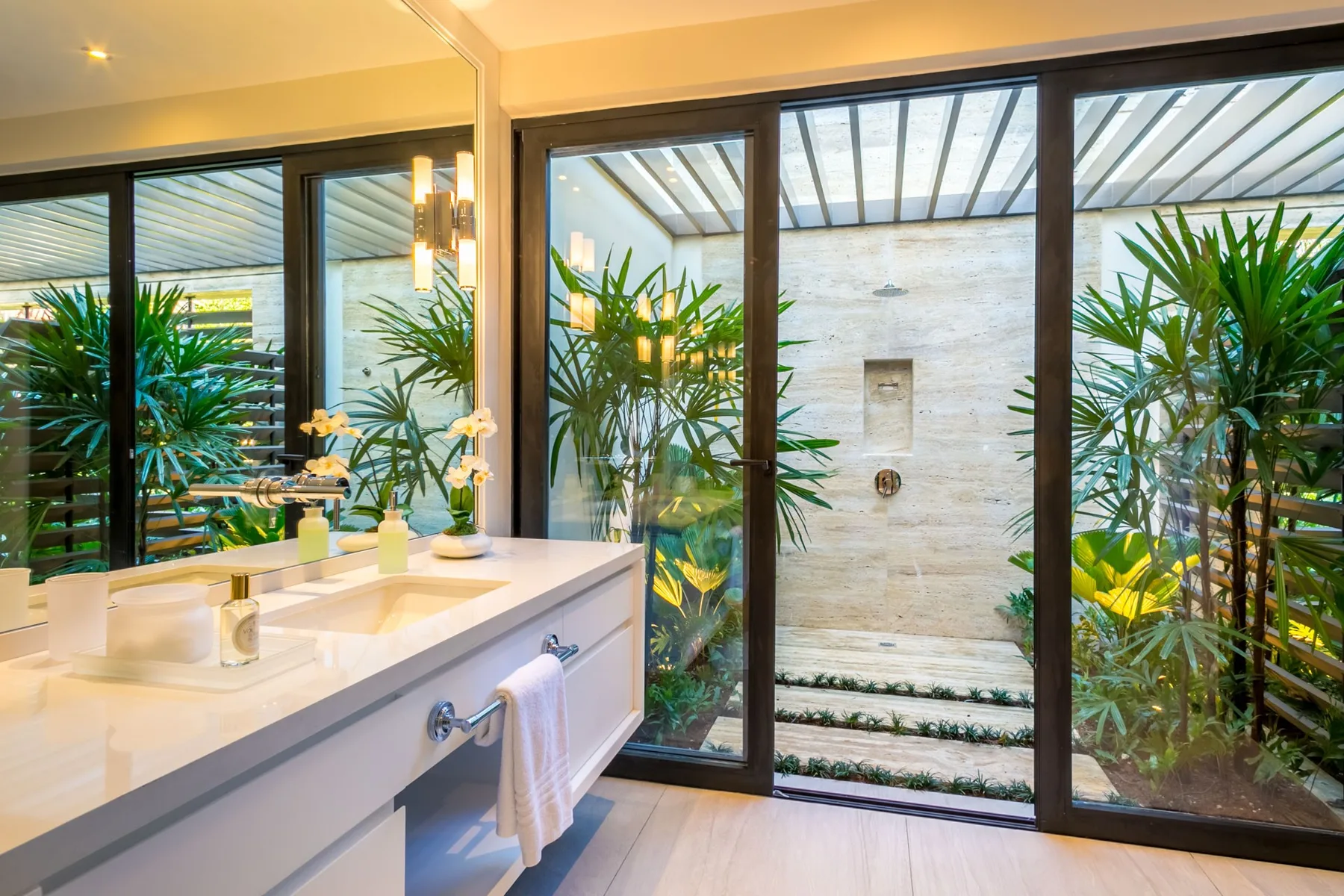A bathroom vanity faces a glass-enclosed shower, with plants visible beyond.