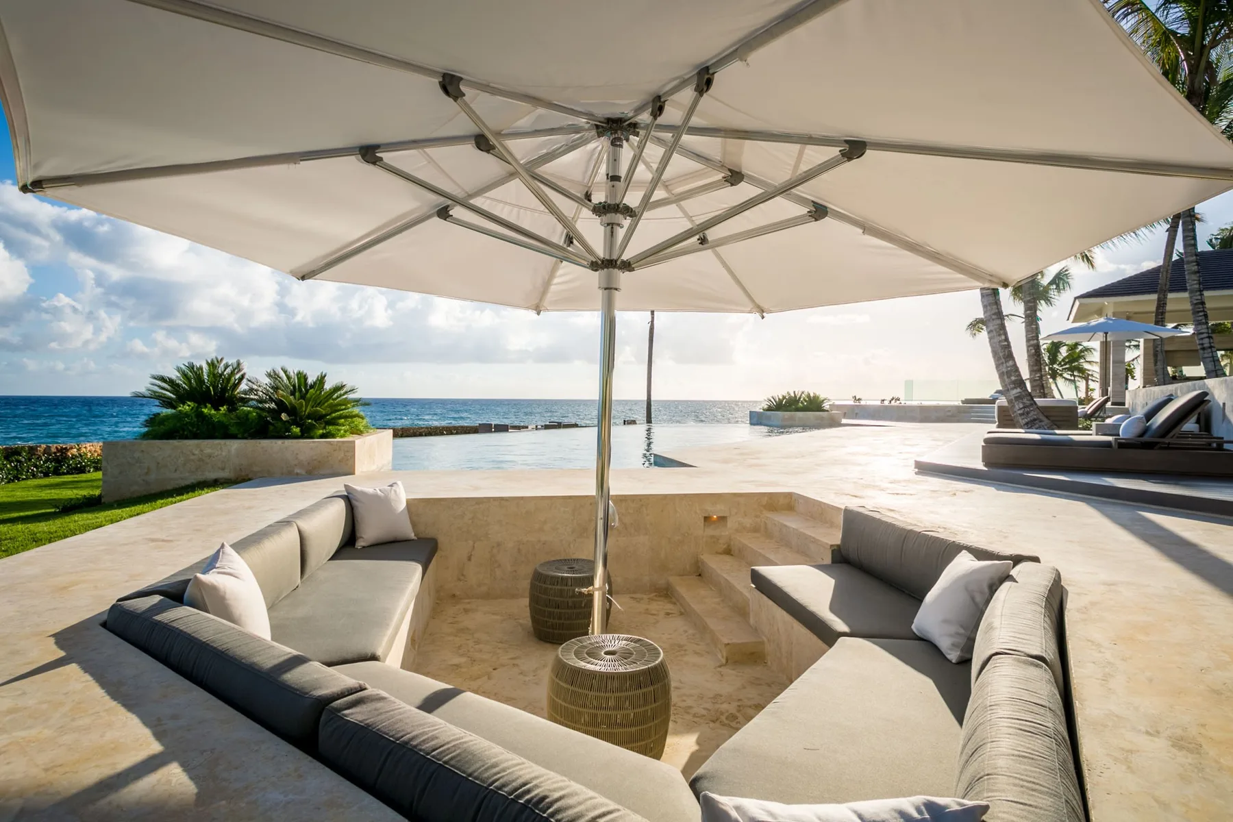 A sunken outdoor seating area sits beneath a large umbrella beside the ocean.