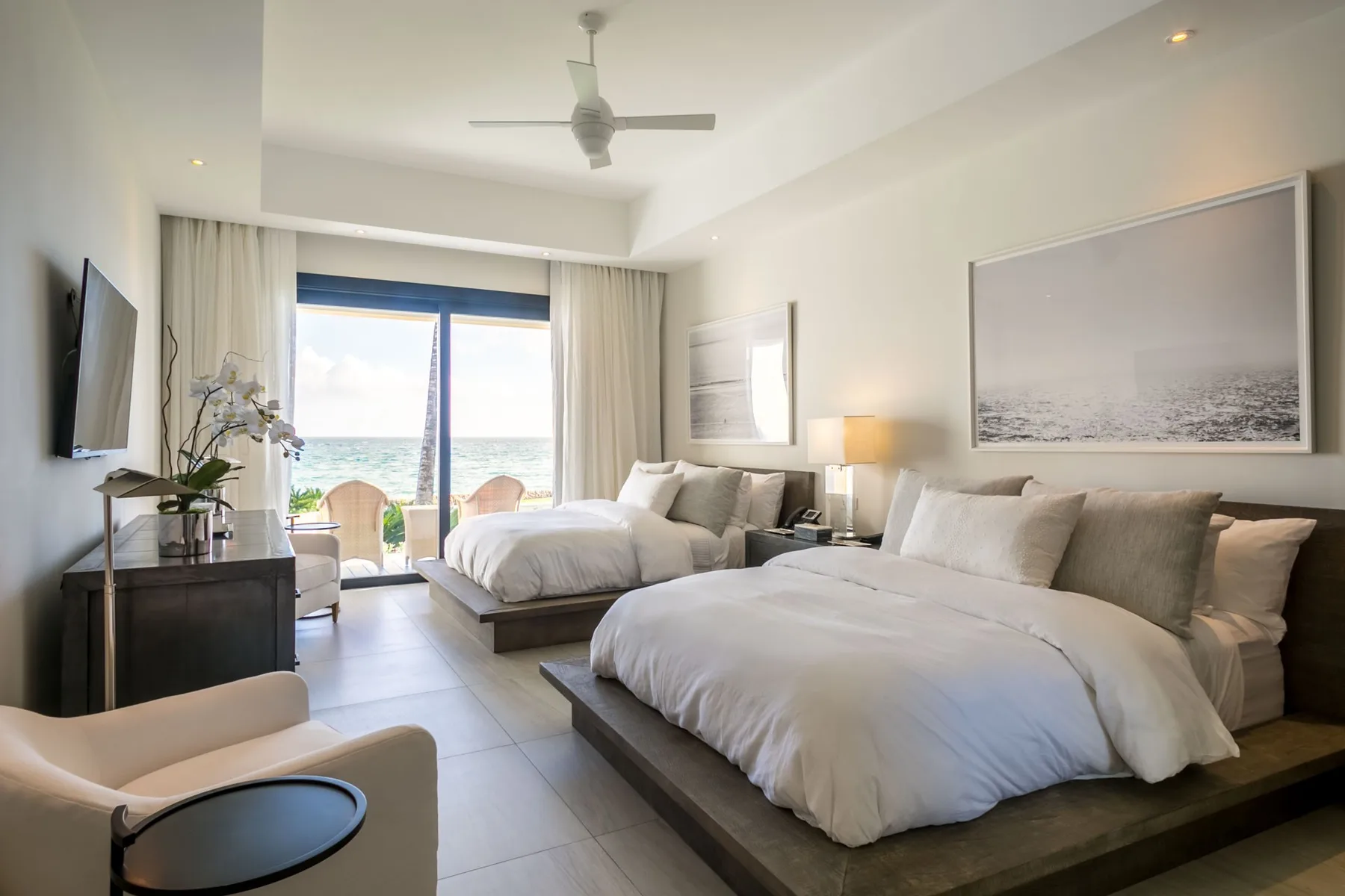 A bright bedroom with two beds, a television, and glass doors opening onto a balcony.
