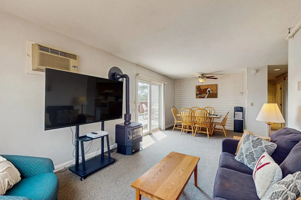 Una sala de estar con asientos, un televisor y una mesa de comedor junto a las ventanas.