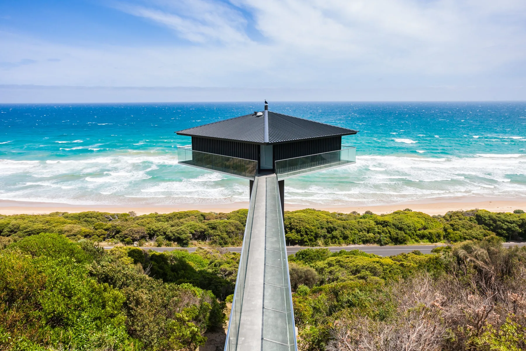 A raised house stands above coastal vegetation, facing the ocean and beach.