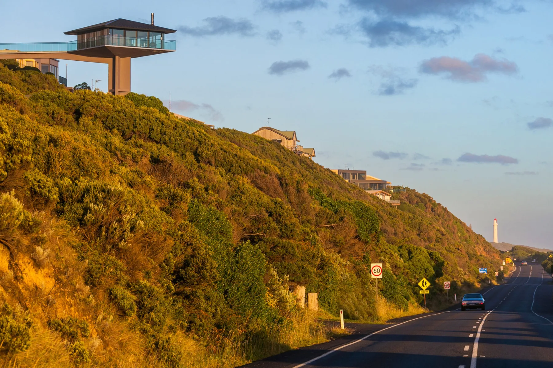 A coastal hillside road runs below a modern house perched above the slope.