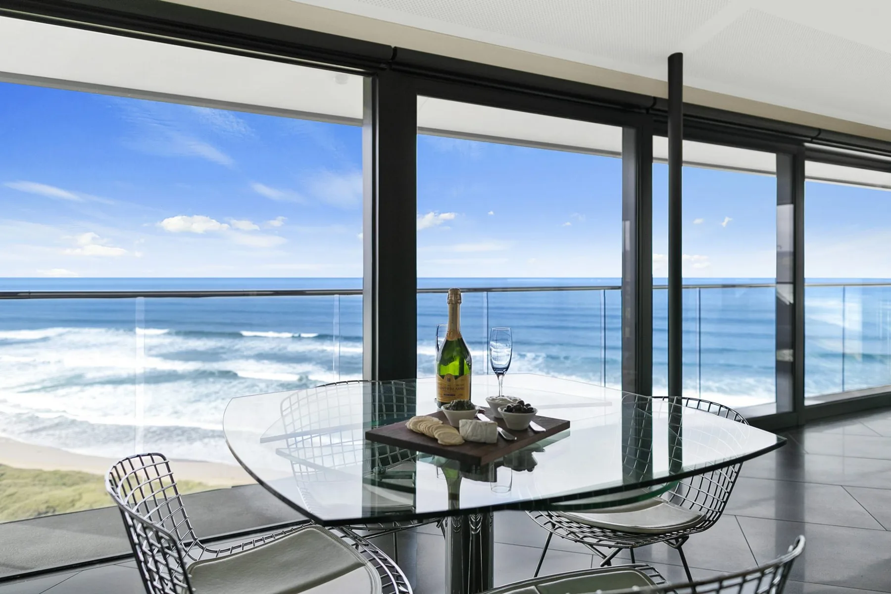 A glass dining table and chairs overlook the ocean through wide windows.