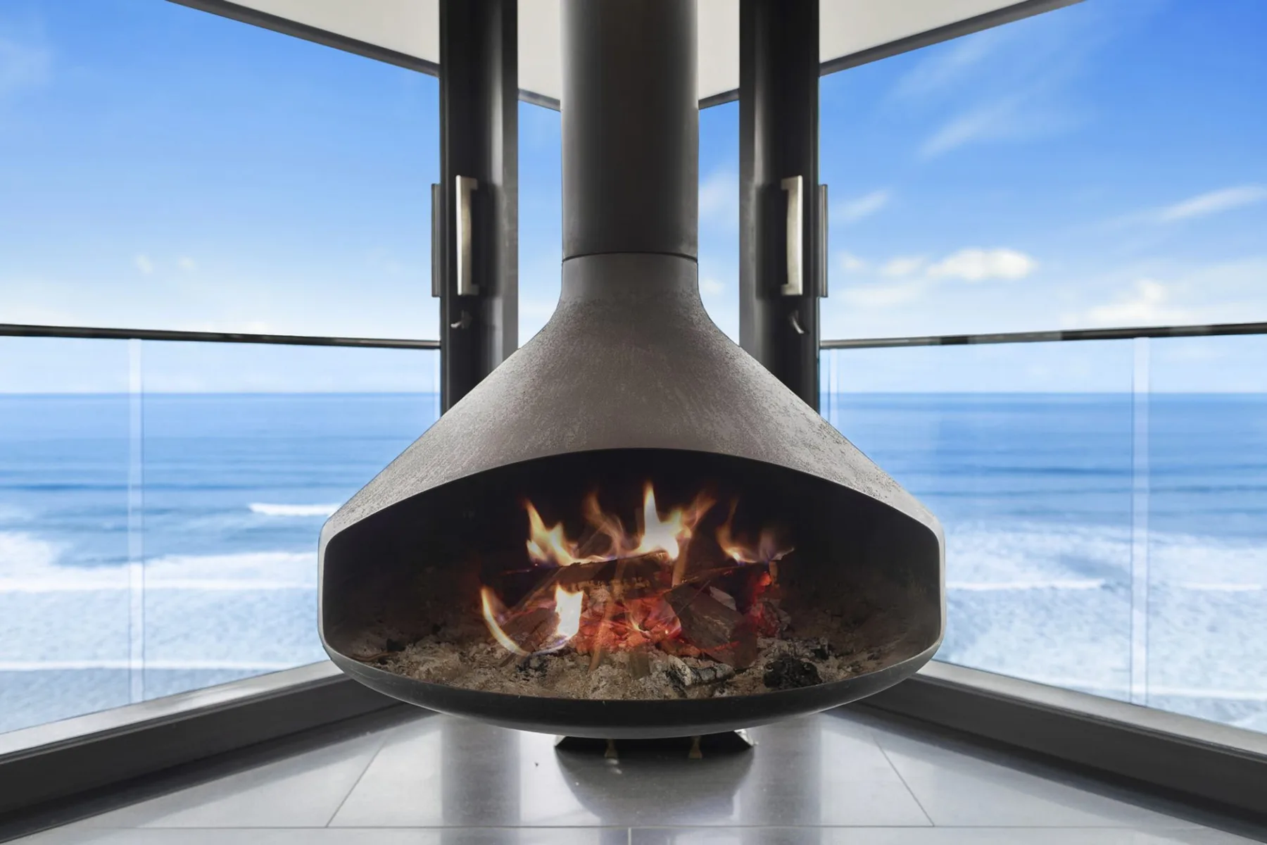 A suspended fireplace burns in front of floor-to-ceiling windows overlooking the ocean.
