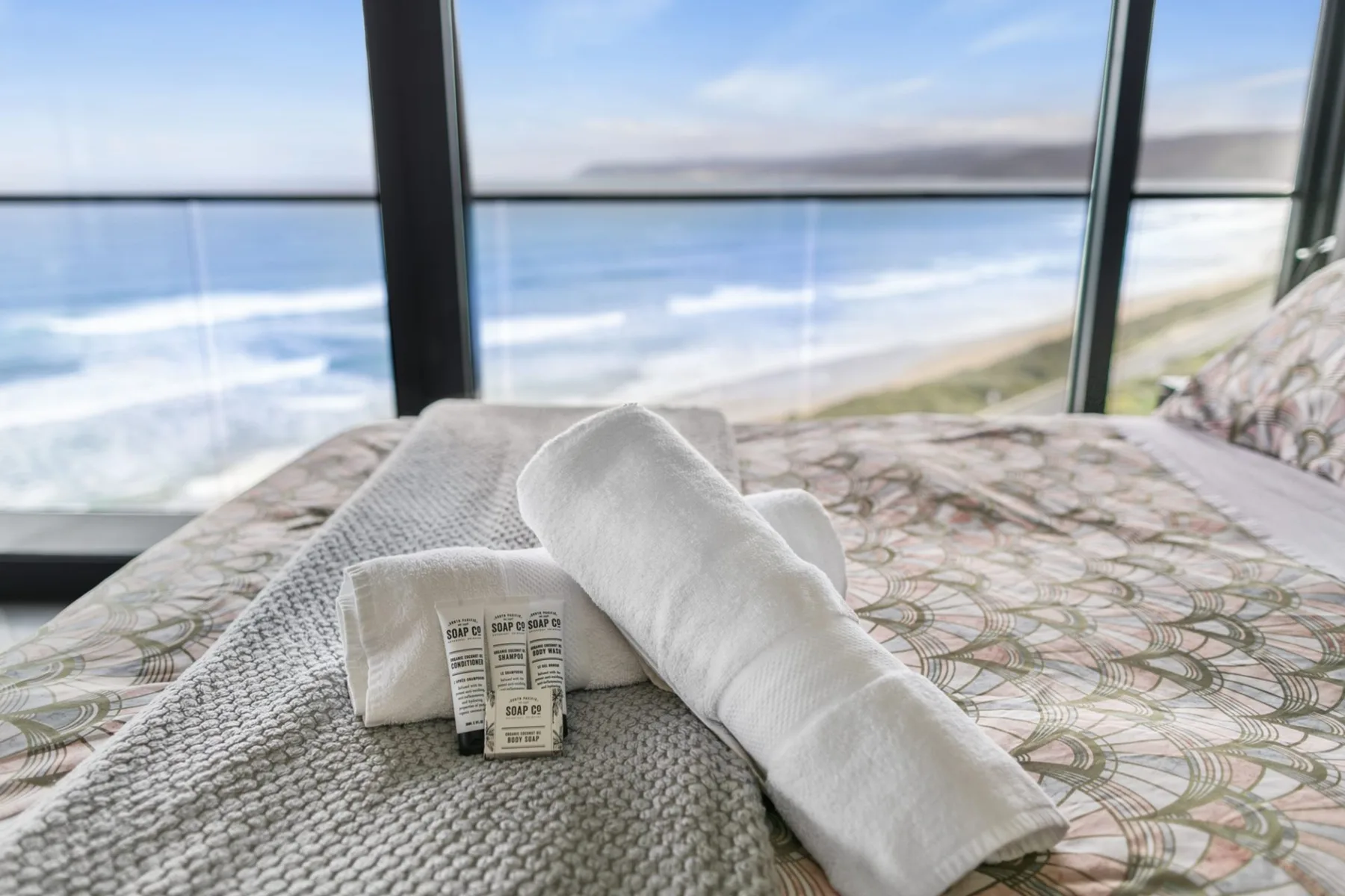 A bedroom with folded towels on the bed beside a large window overlooking the ocean.