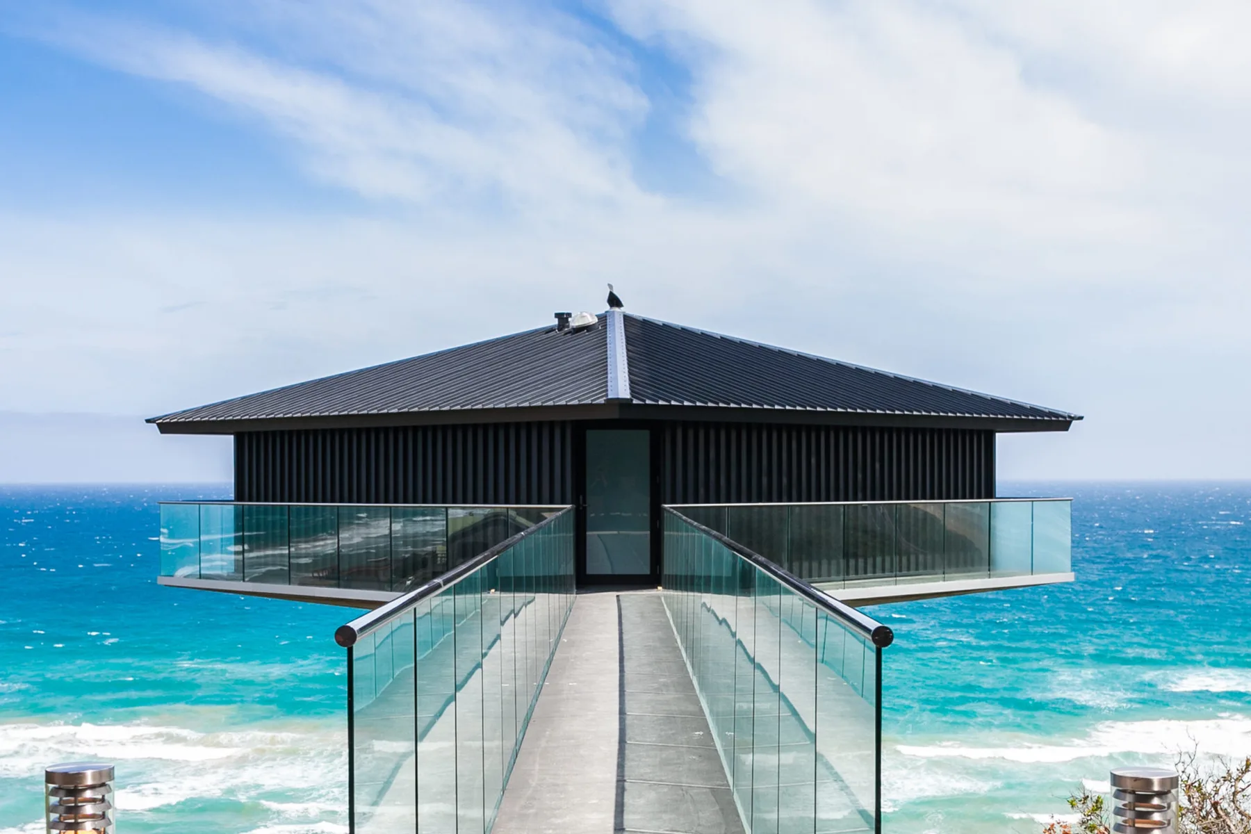 A modern house stands over the ocean at the end of a narrow walkway.