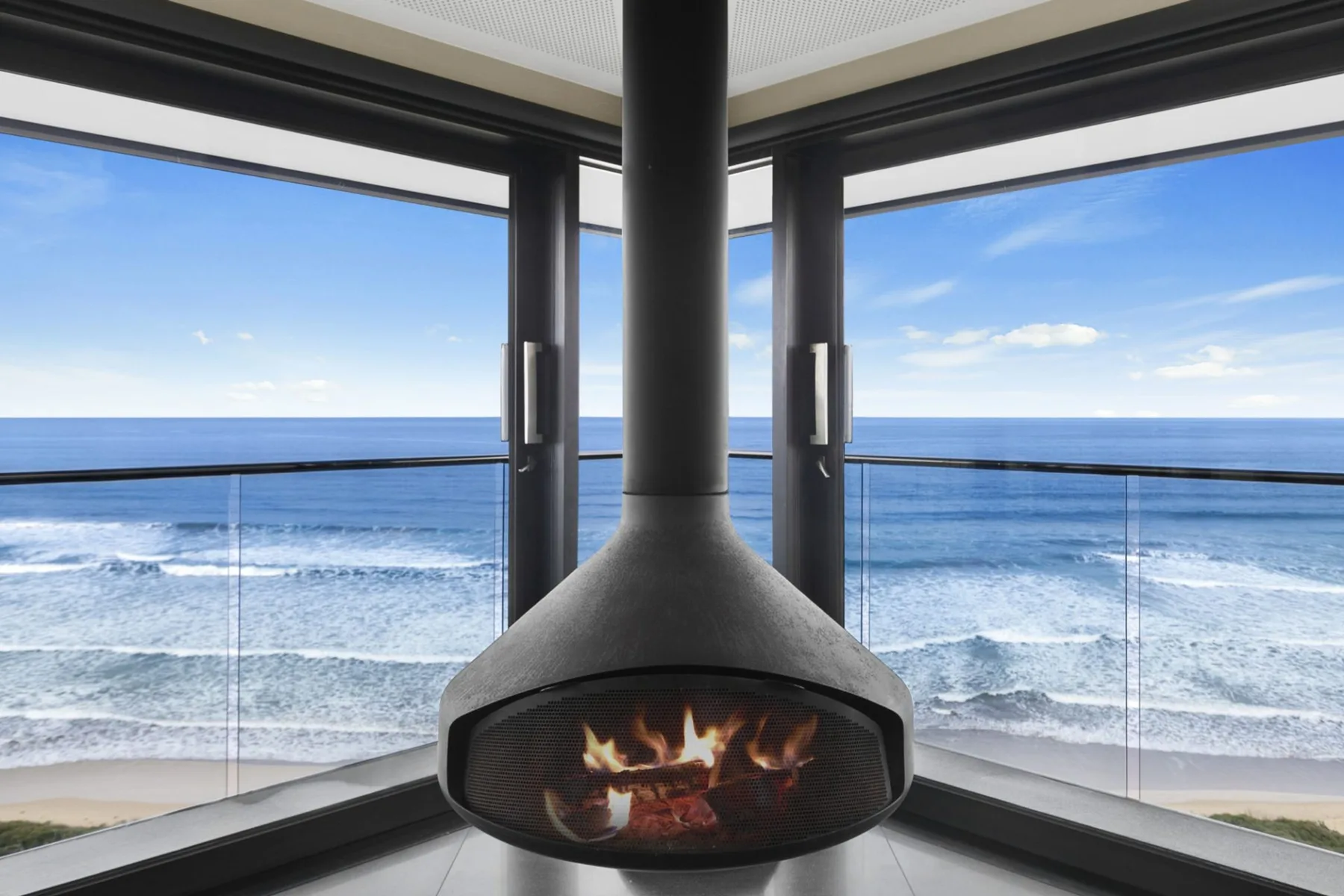 A room with a suspended fireplace overlooking the ocean through large windows.