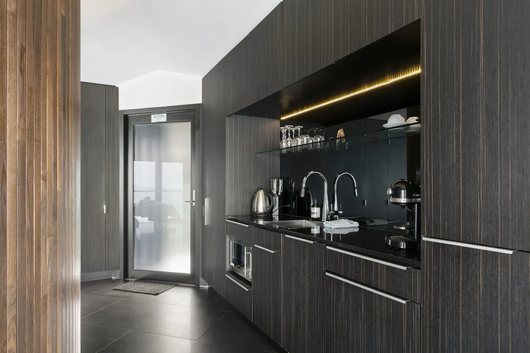 A dark modern kitchen with a glass-fronted refrigerator and countertop appliances.
