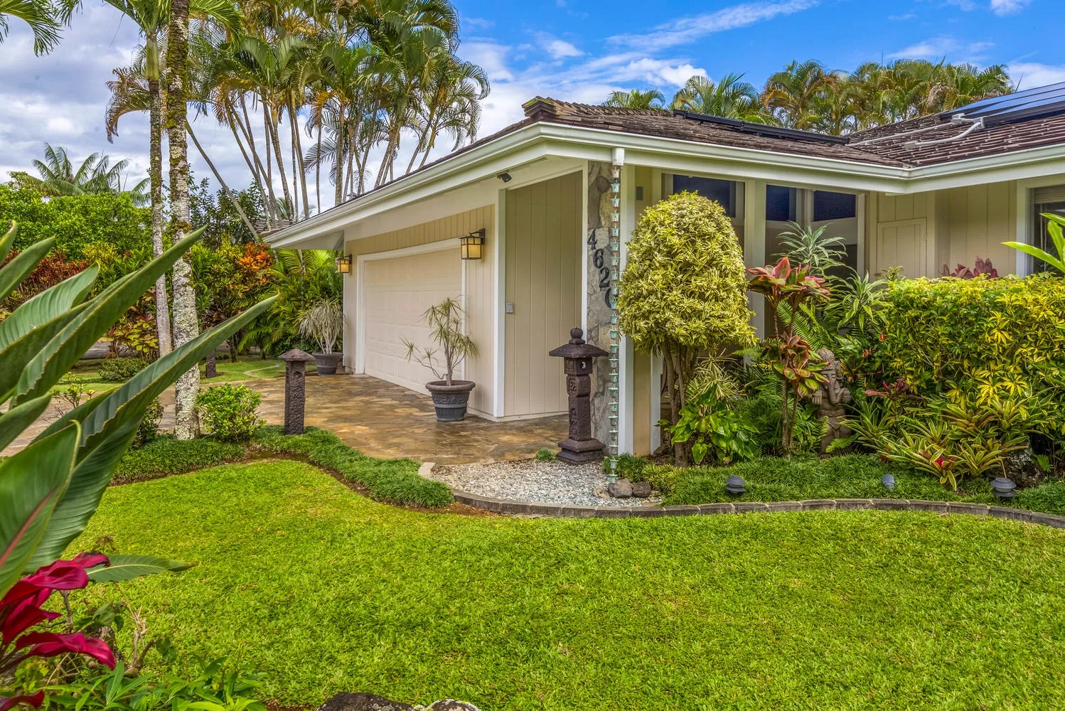 A single-story house stands behind a green lawn, with tropical plants around the entrance.