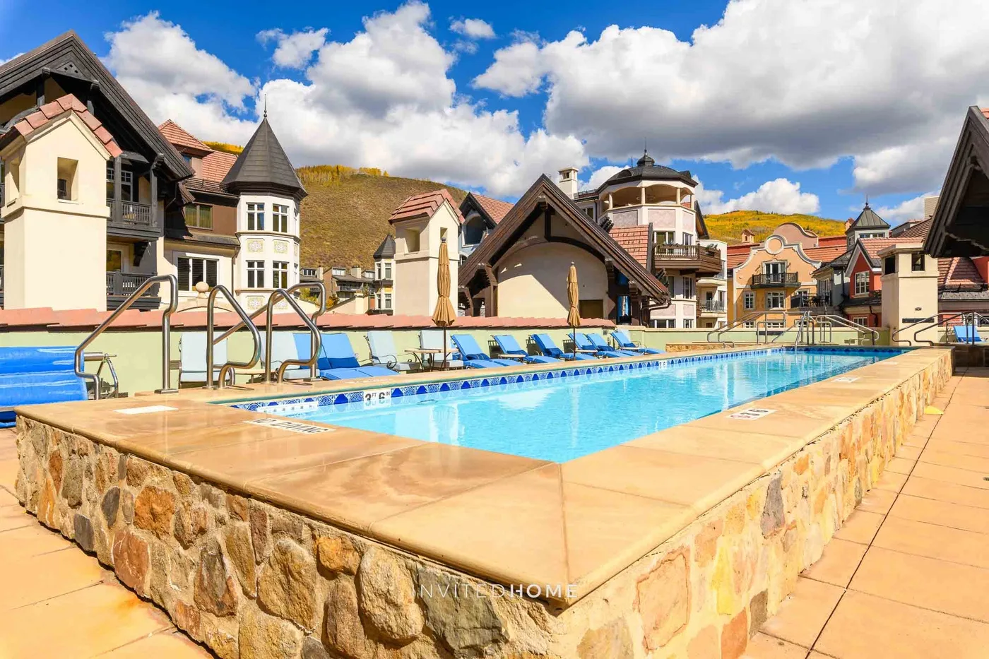Una piscina al aire libre está rodeada de edificios de estilo alpino bajo un cielo parcialmente nublado.