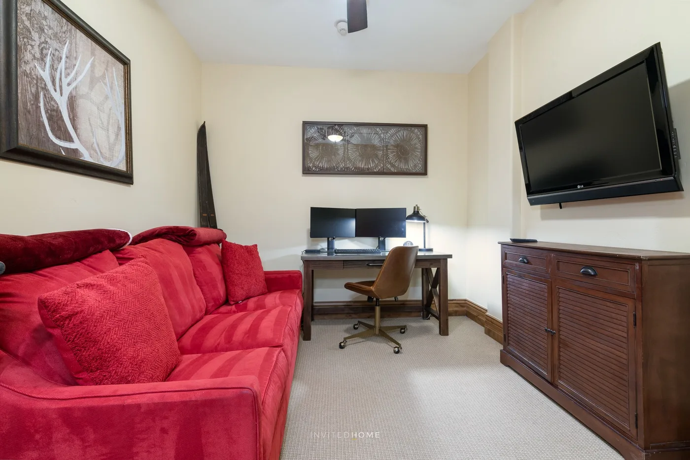 Una oficina en casa con escritorio y silla junto a un sofá rojo y un televisor en la pared.