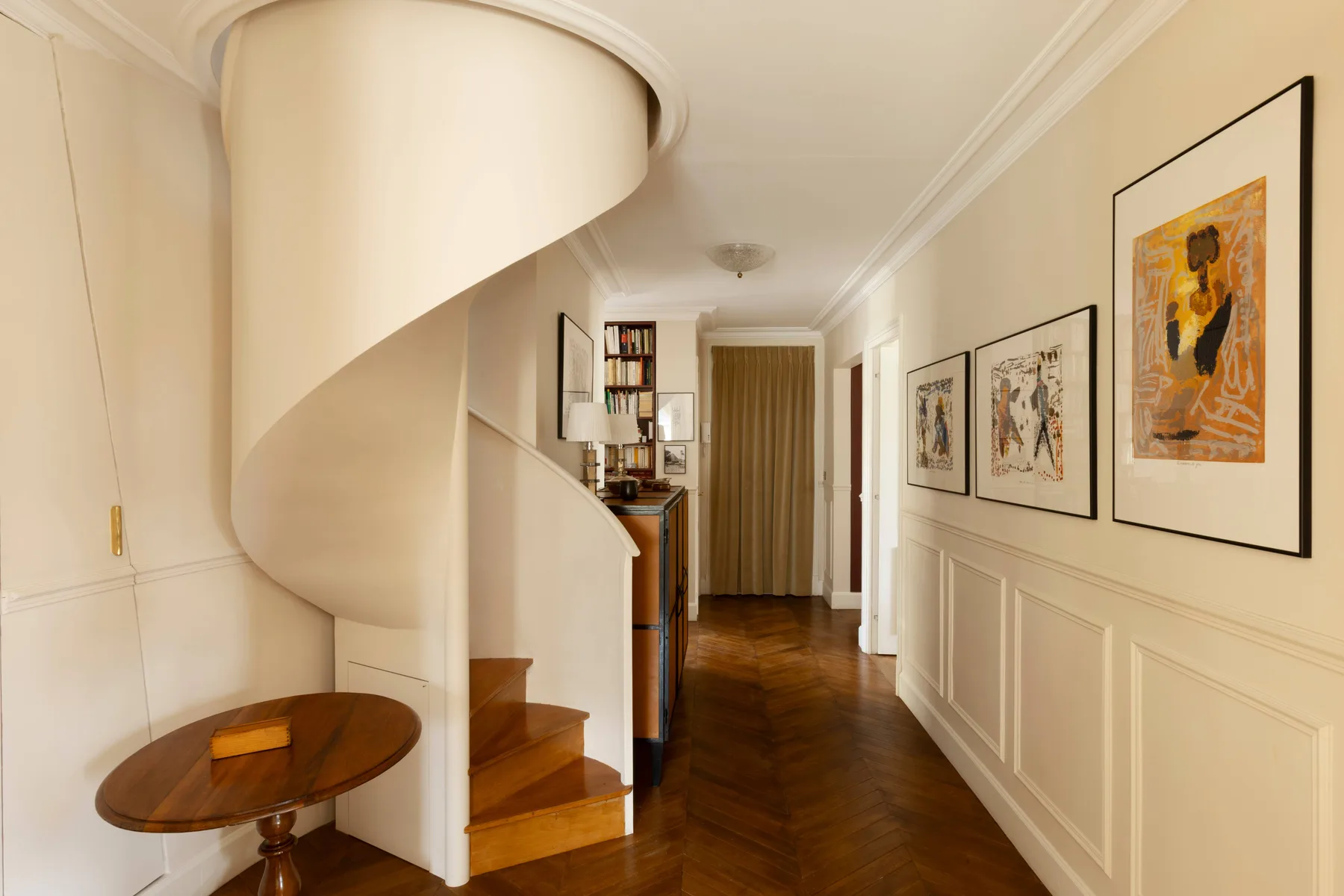 Un pasillo con una escalera curva, cuadros enmarcados y suelo de madera oscura.