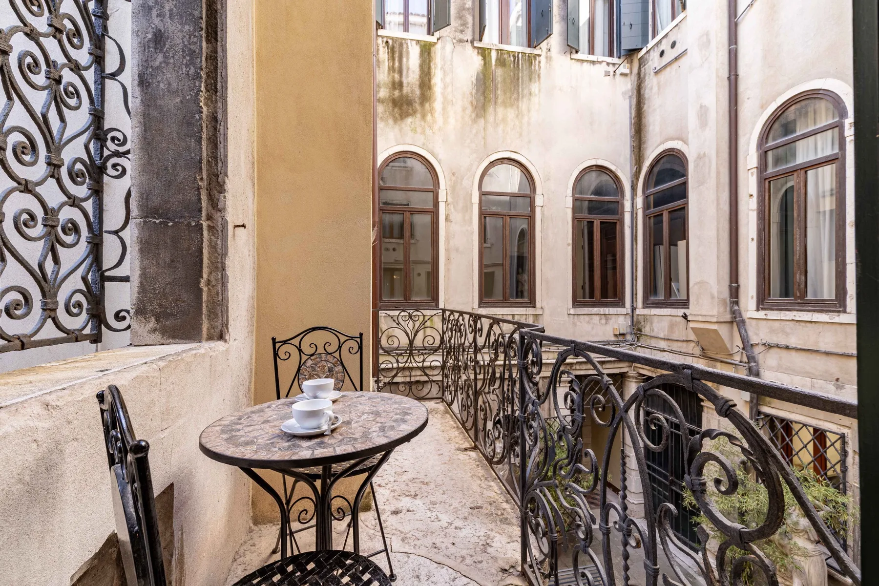 A small balcony with a round table and ornate metal railings overlooks an interior courtyard.
