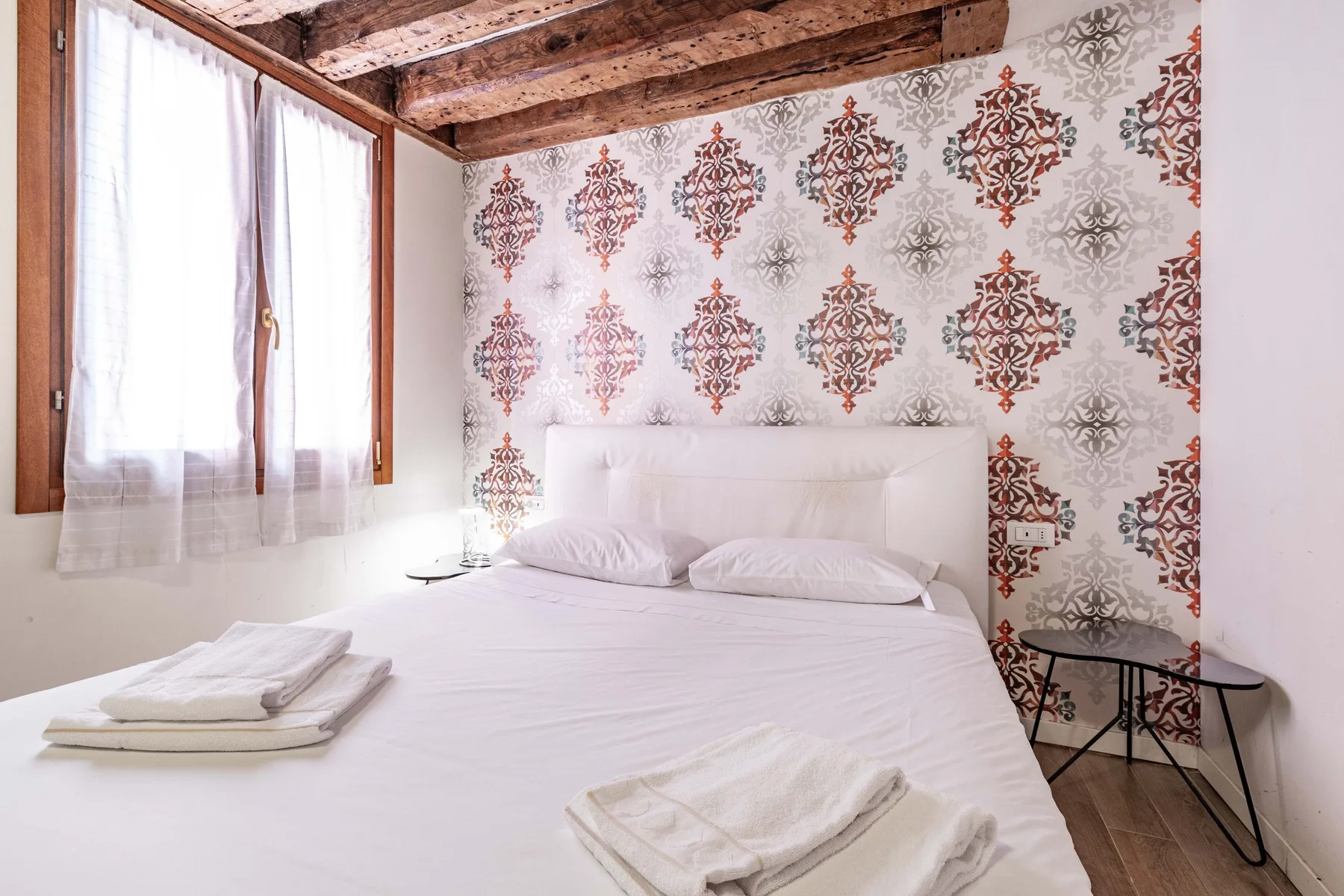 A bedroom with a bed, patterned wallpaper, and a window with sheer curtains.