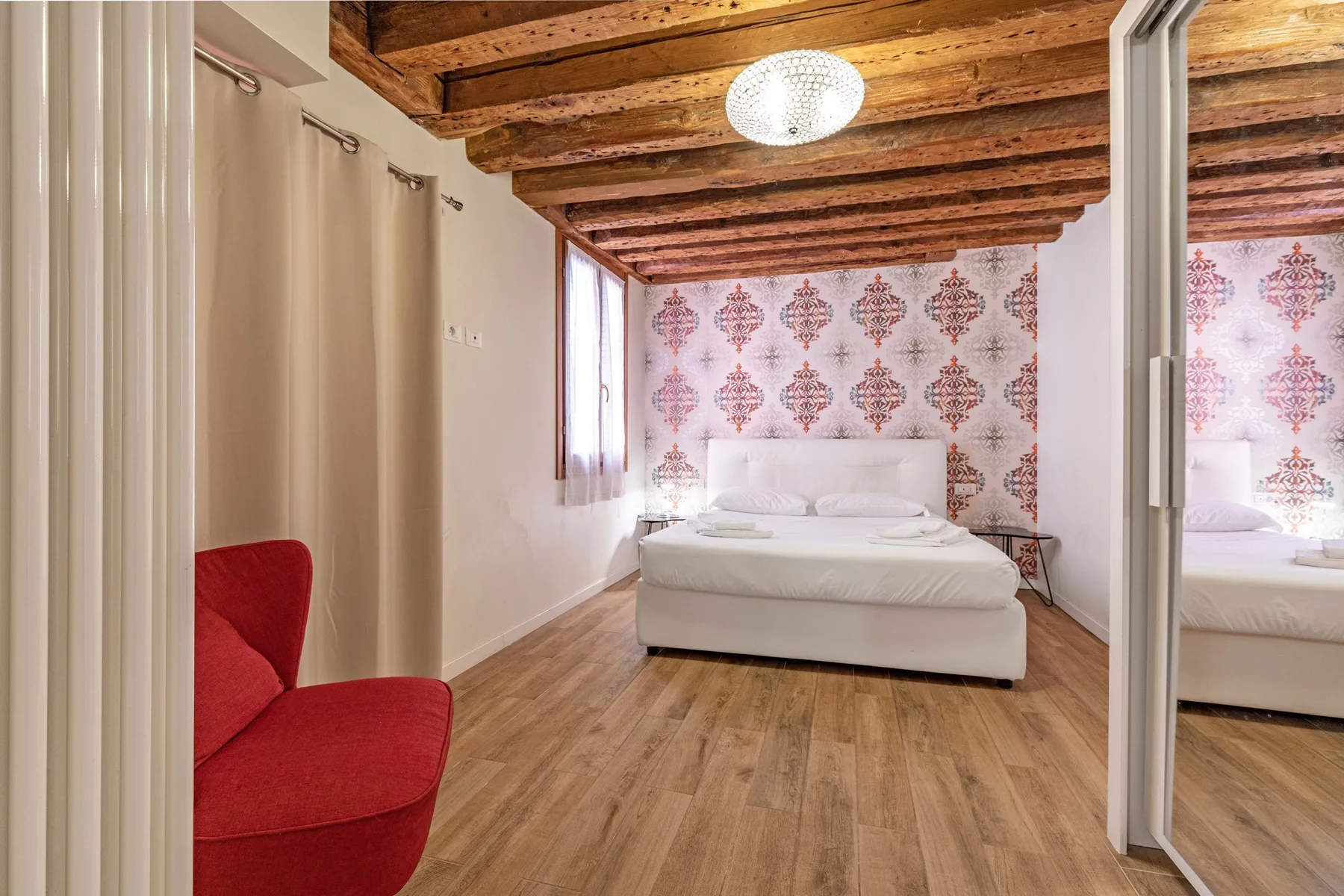 A bedroom with a white bed, patterned wallpaper, and exposed wooden ceiling beams.