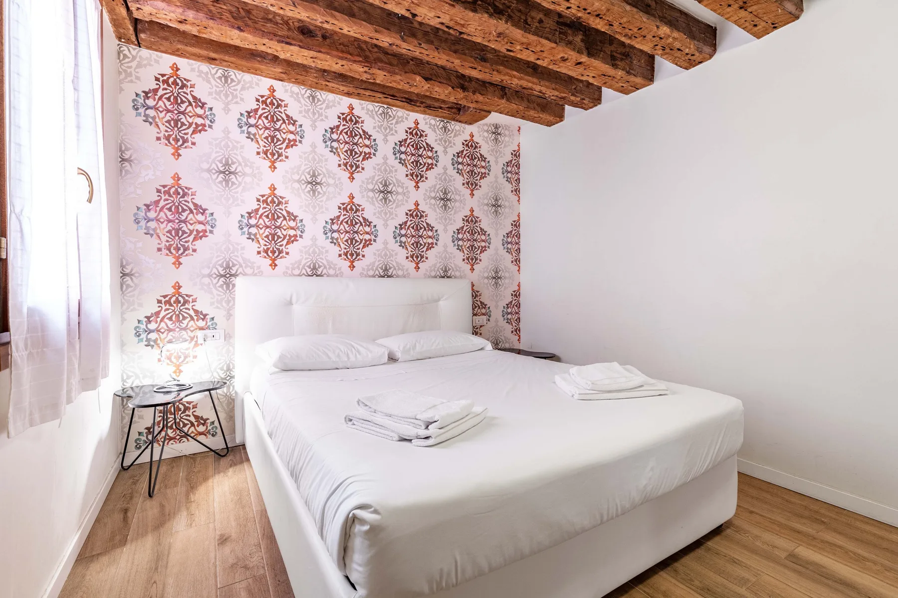 A bedroom with a white bed, patterned wall, and exposed wooden ceiling beams.