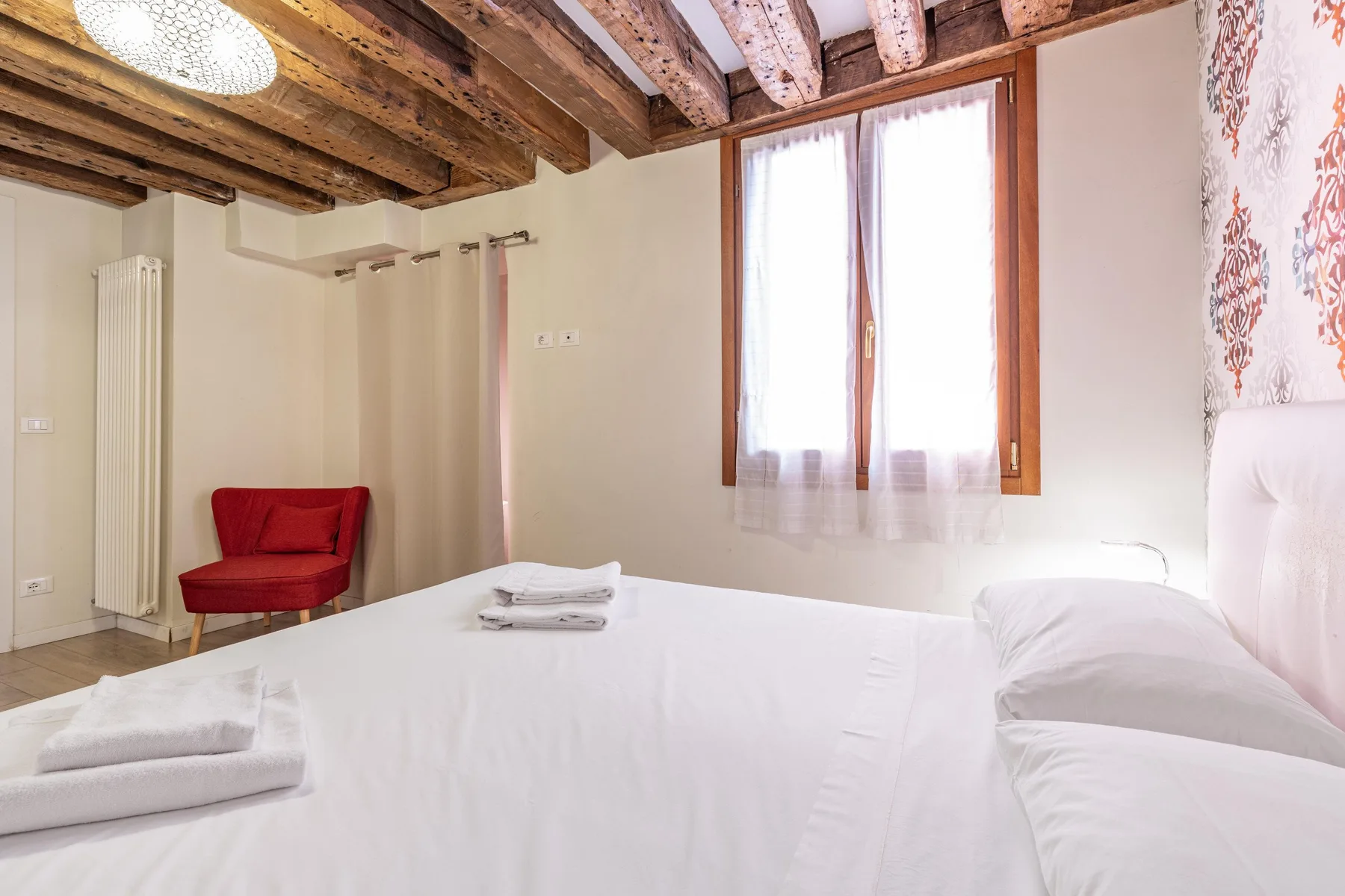 A bright bedroom with white bedding, a red chair, and a wooden-beam ceiling.
