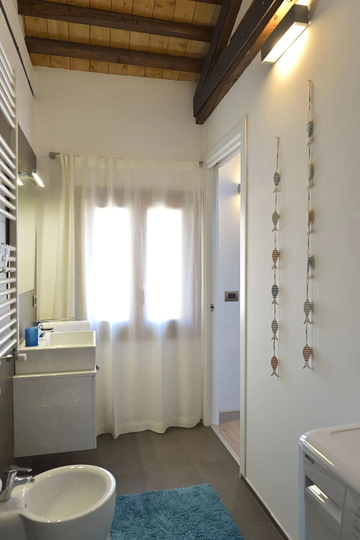 Un baño luminoso con lavabo, cortinas translúcidas y vigas de madera a la vista.