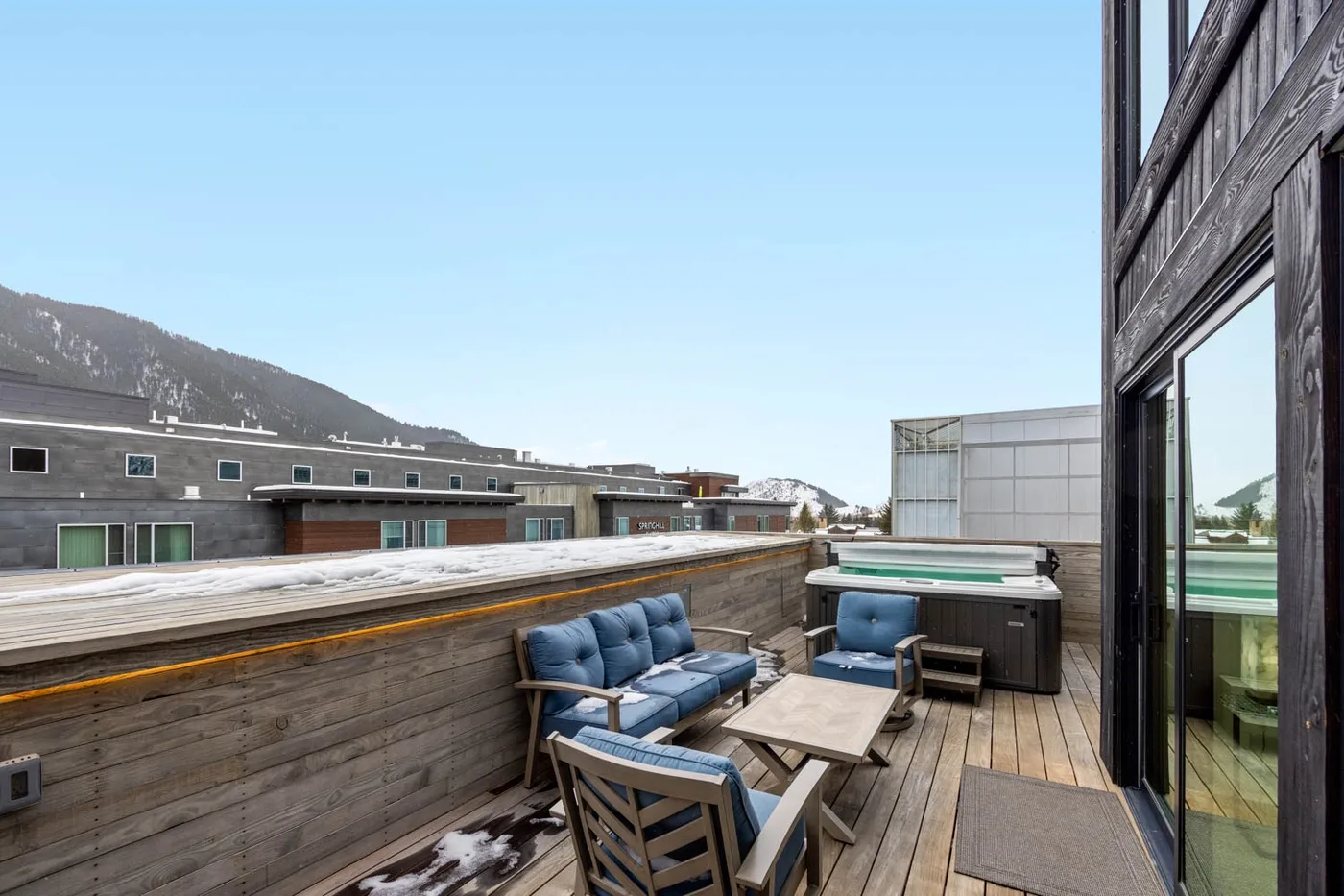 A wooden deck with outdoor seating and a hot tub overlooks nearby buildings and mountains.