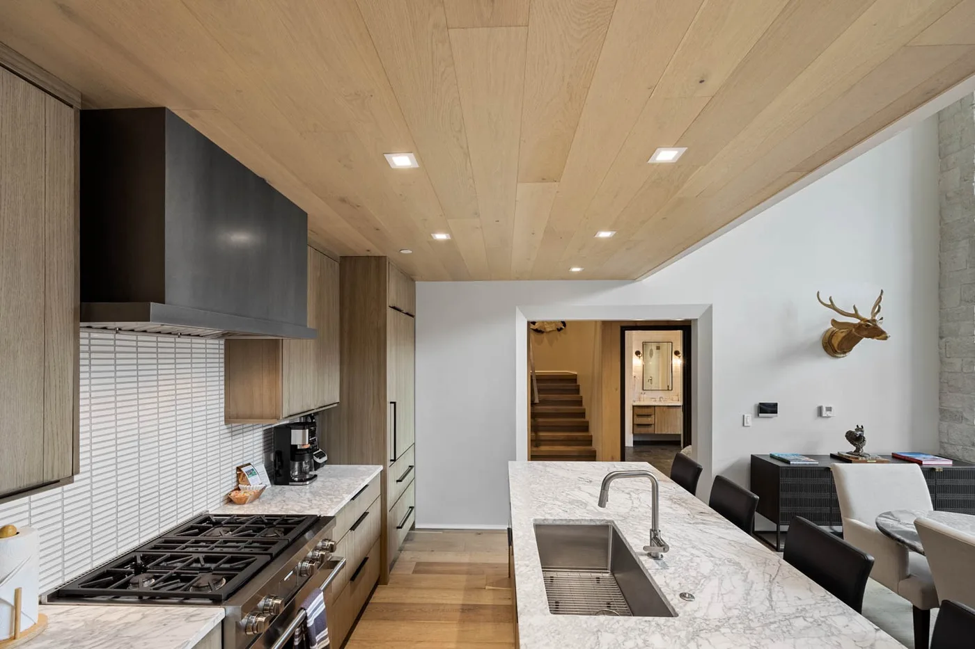 A modern kitchen opens onto a dining area, with a central island and staircase beyond.