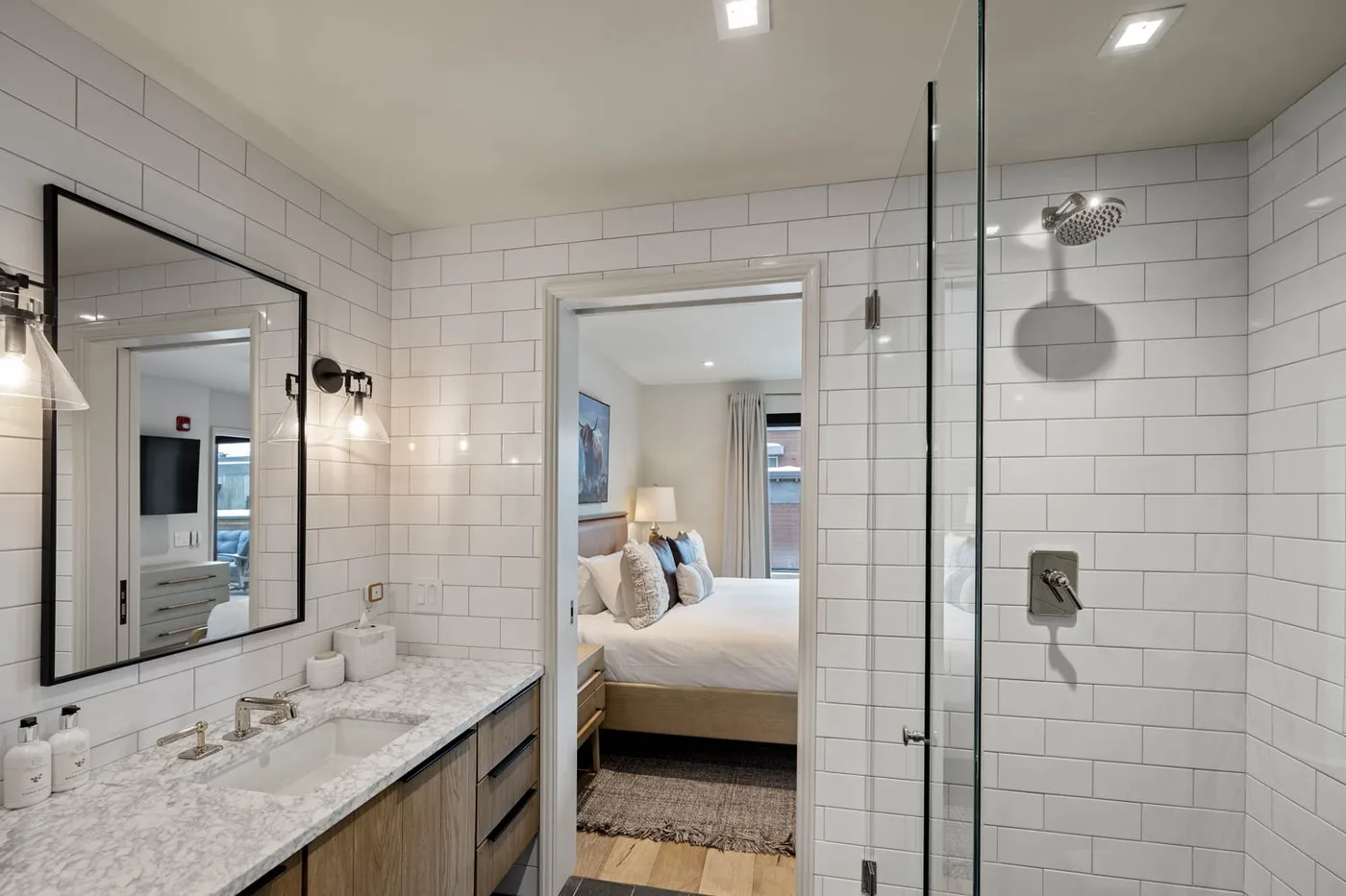 A tiled bathroom with a glass shower, vanity, and an open doorway to a bedroom.