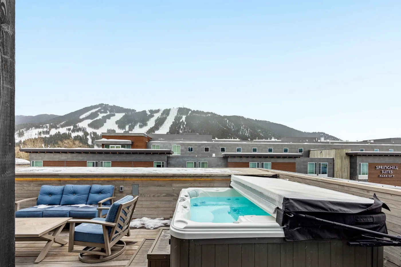 A rooftop deck with a hot tub and outdoor seating overlooks a mountain slope.