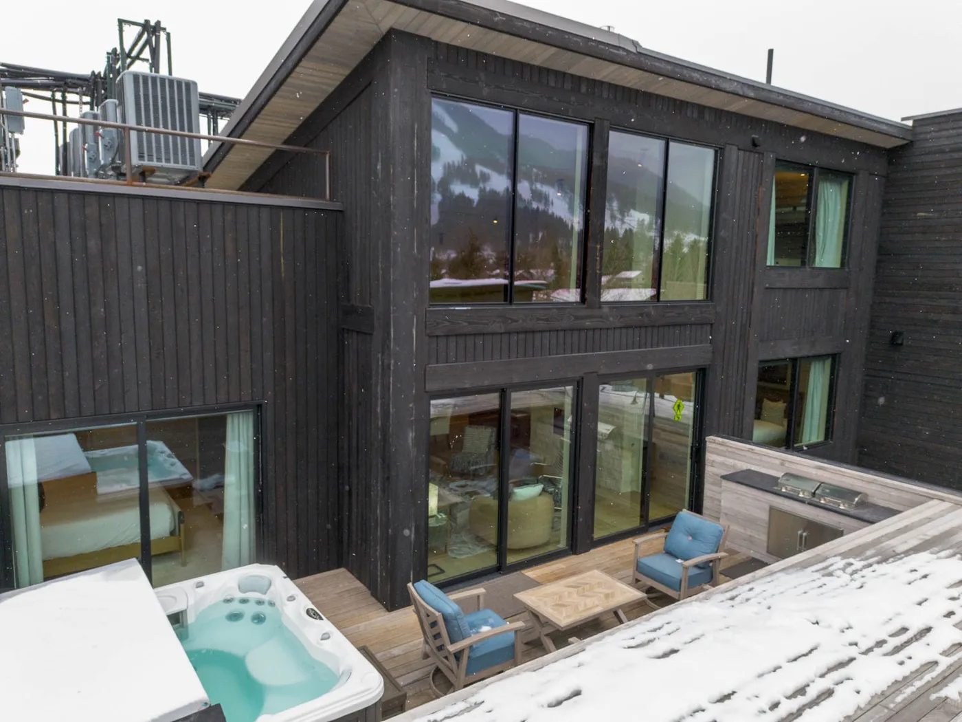 A dark-sided two-story building with large windows opens onto a wooden deck with seating and a hot tub.