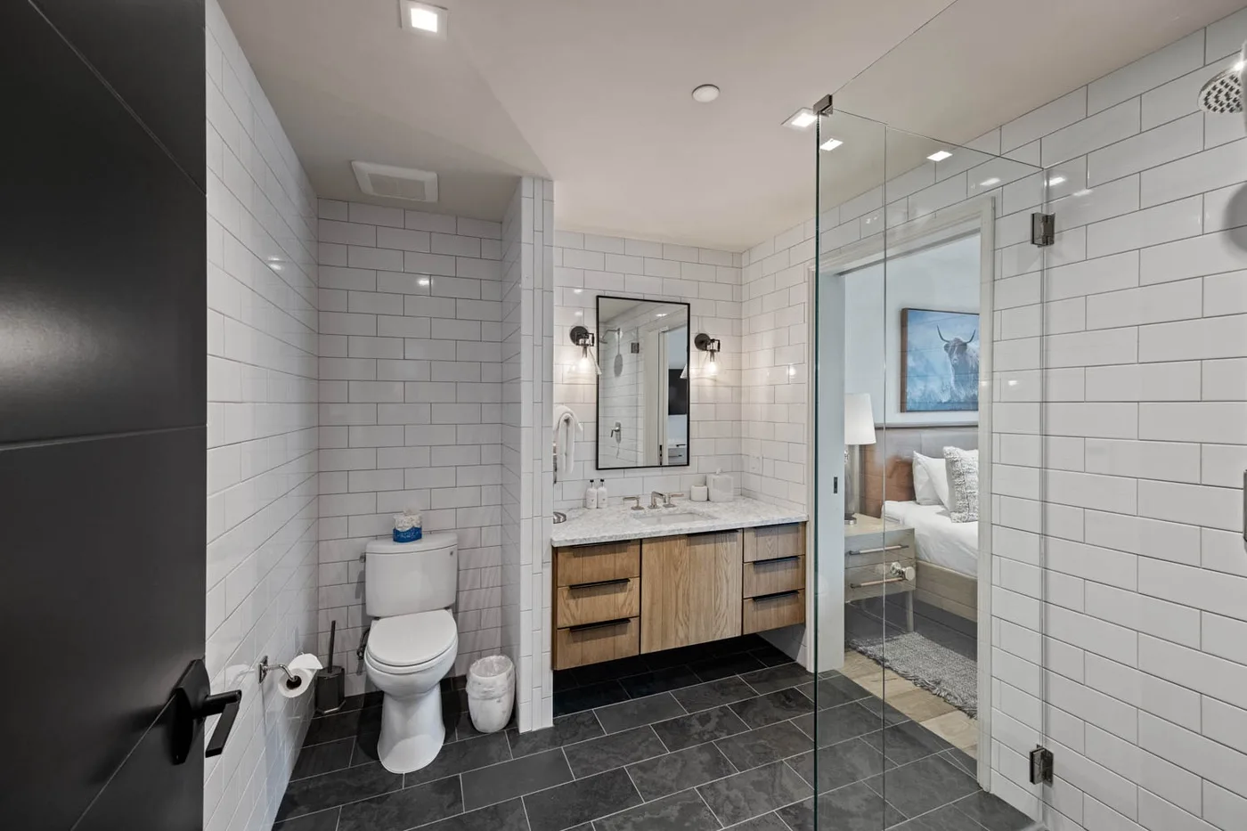 A tiled bathroom with a vanity, toilet, and glass shower beside a bedroom.