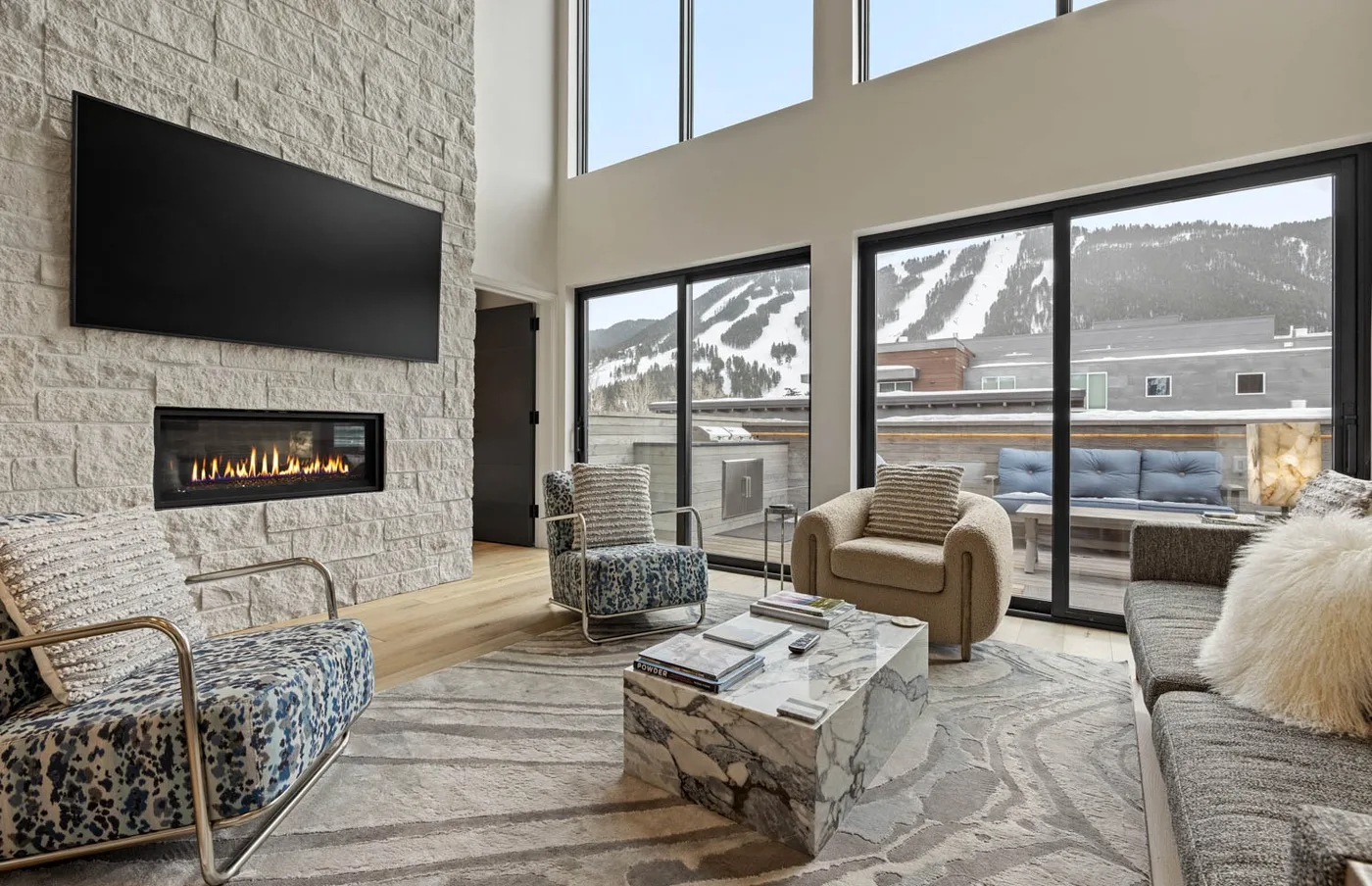 A bright living room with a fireplace, seating, and large windows overlooking a snowy mountain.