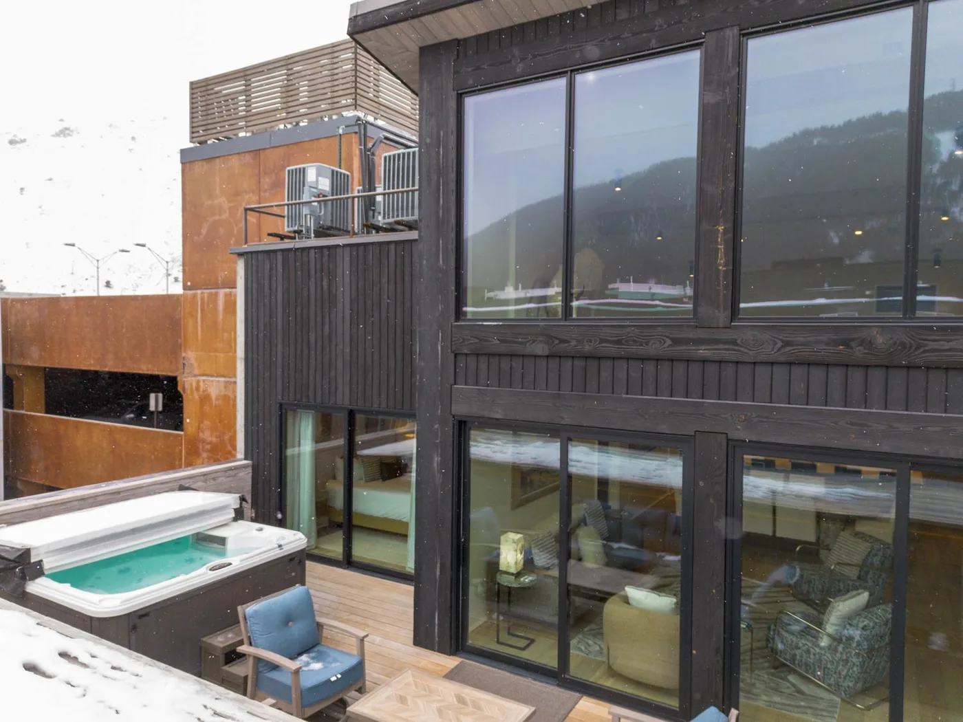 A modern building facade opens onto a snowy terrace with a hot tub and seating.