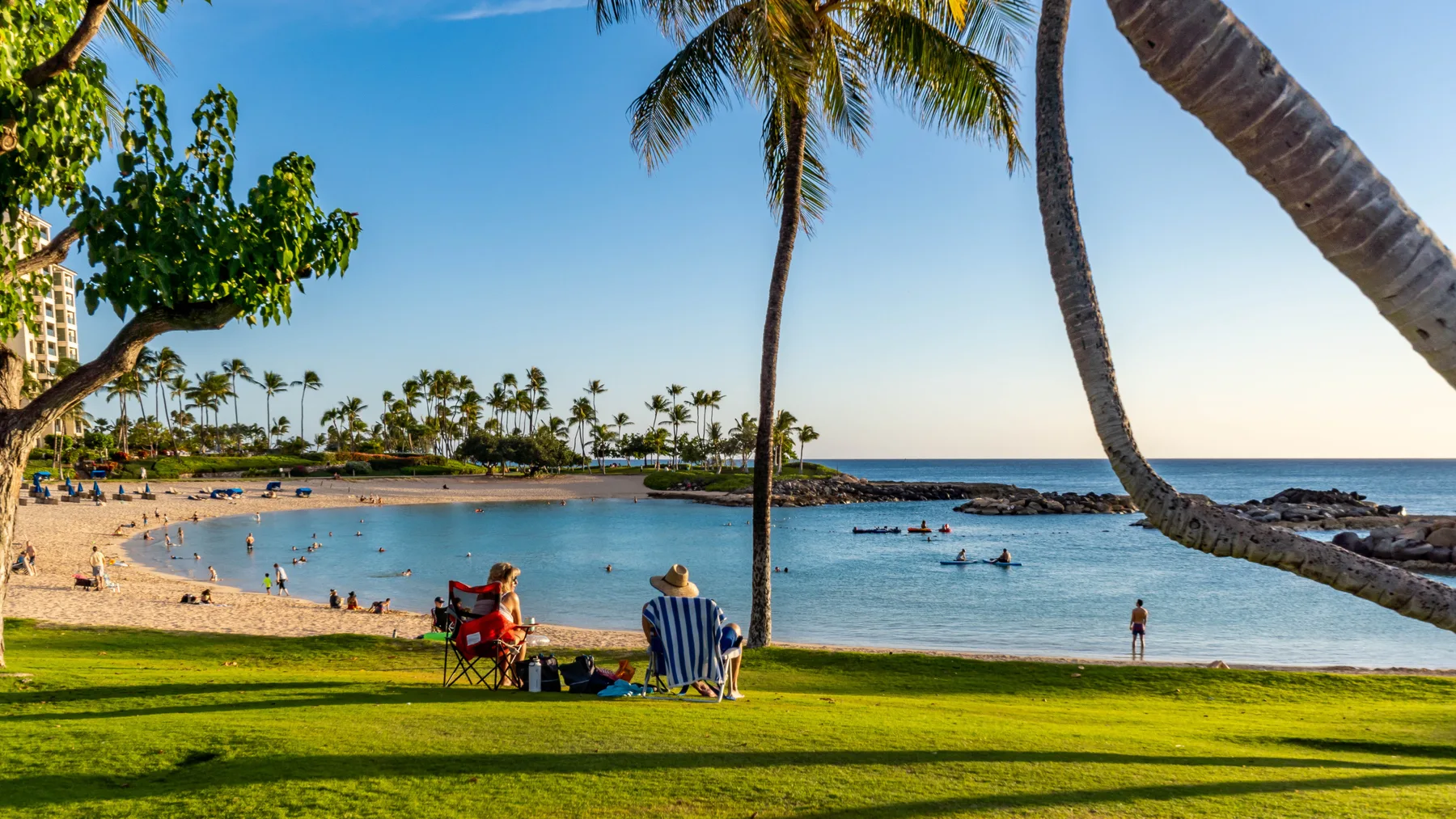 A tropical beach with calm water, palm trees, and people relaxing along the shore.
