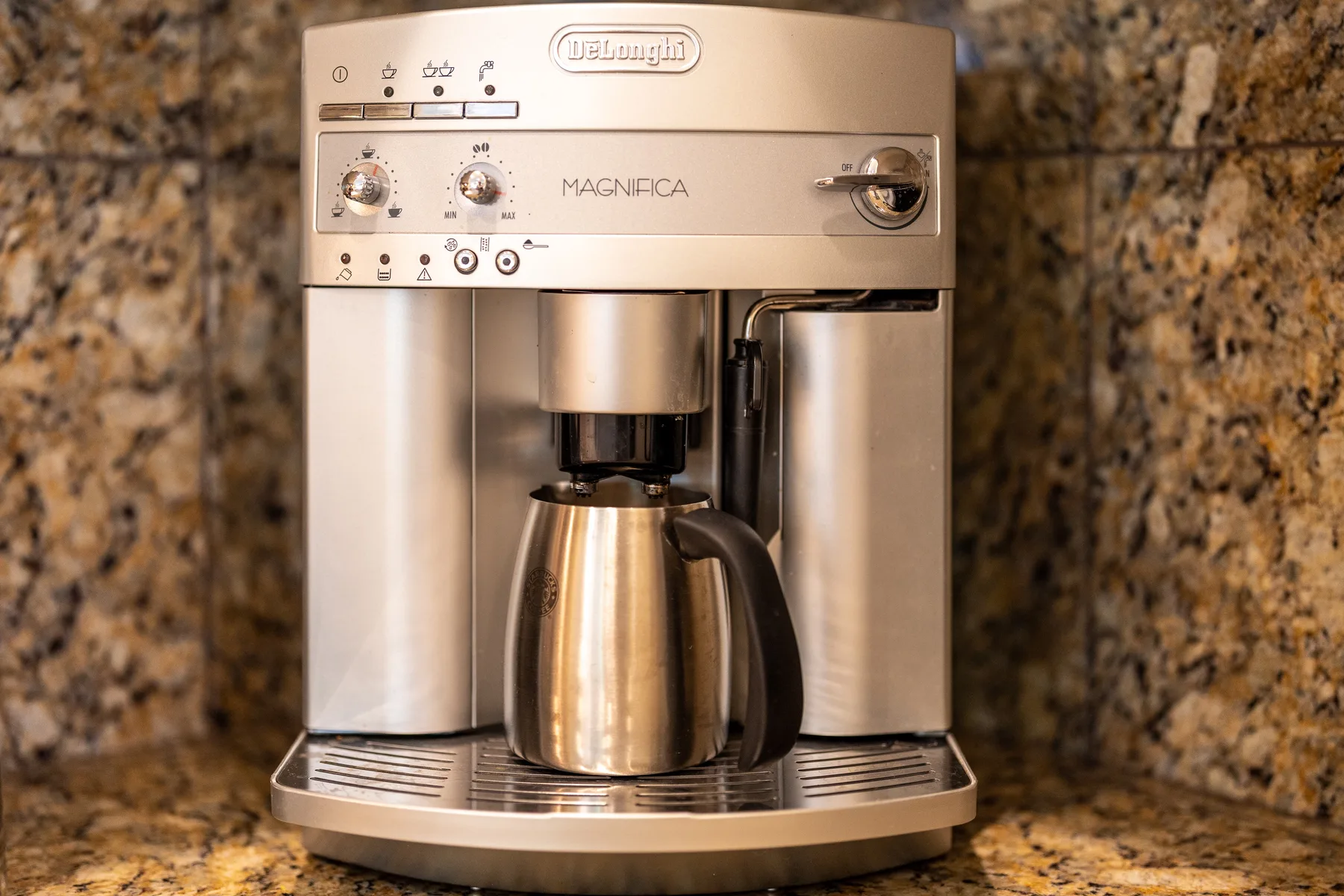 A countertop coffee machine with a metal carafe sits against a stone-patterned wall.