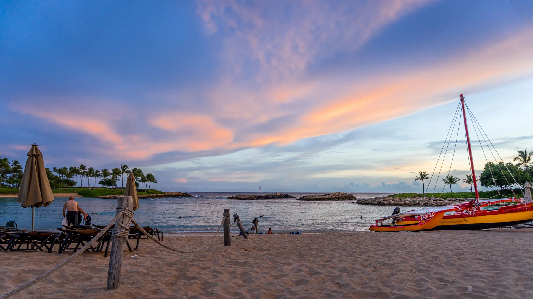 A sandy beach beside calm water, with lounge seating and colorful sailboats at sunset.