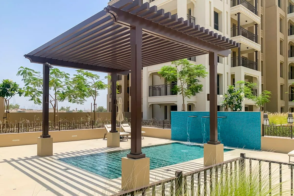 Una piscina exterior está bajo una pérgola sombreada junto a un edificio de apartamentos.