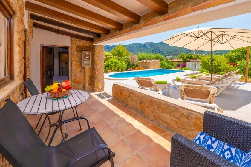 A covered patio with outdoor seating overlooks a swimming pool and green hills.